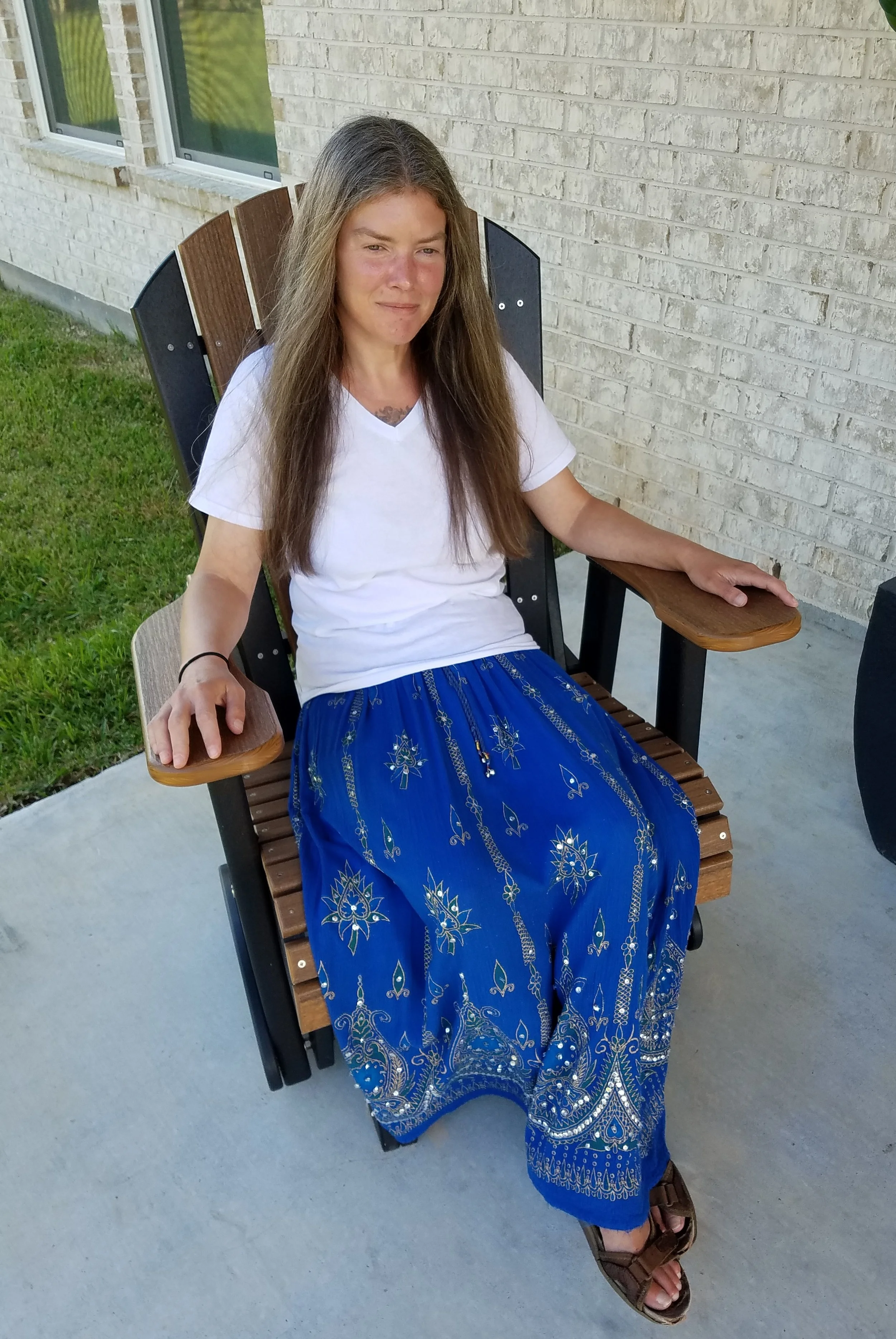 A young woman sitting on a wooden patio chair with a curved backrest, wearing a white t-shirt and a long blue skirt with gold embroidery. She has long brown hair and is sitting outside near a brick wall and windows, with green grass nearby.