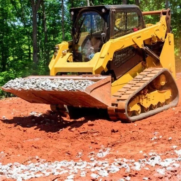 Yellow compact bulldozer on red dirt ground with rocks in the scoop, surrounded by green trees.