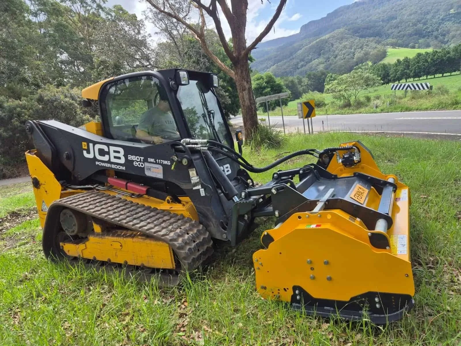 A yellow and black JCB compact track loader with a brush cutter attachment on a grassy roadside next to a highway, with trees and mountains in the background.