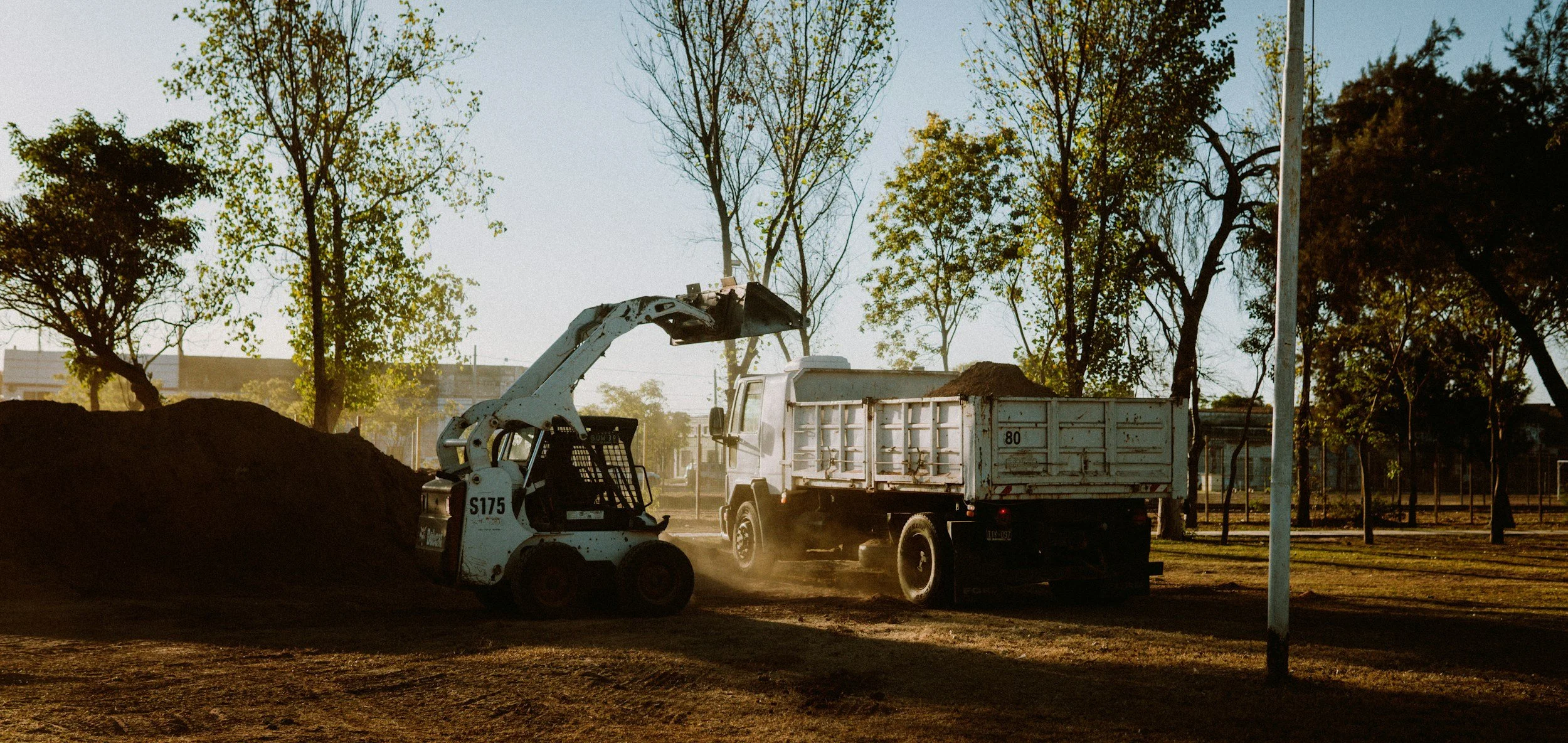 A small construction vehicle loading dirt into a dump truck at a construction site with trees in the background during daytime.