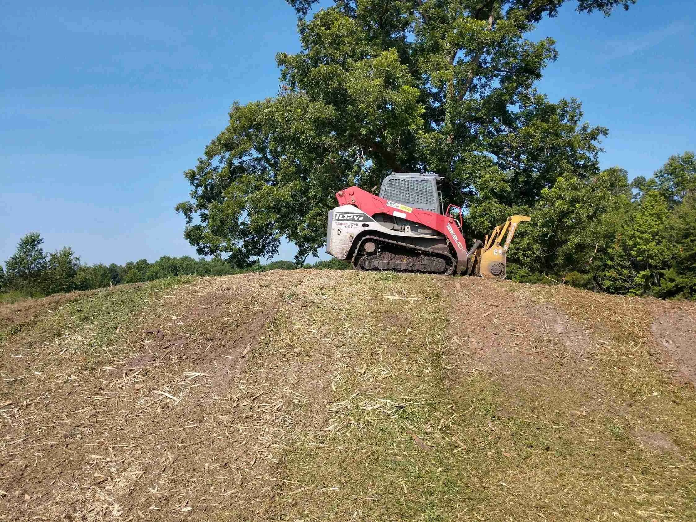A small excavator on a dirt hill removing soil with trees and a clear blue sky in the background.