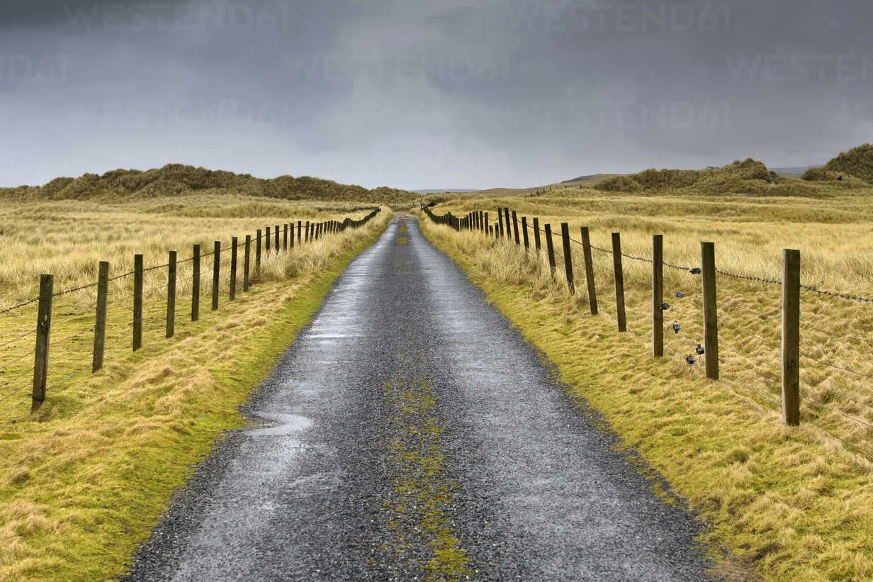 A long, straight gravel road running through a grassy field with wooden fences on both sides and hills in the background under an overcast sky.