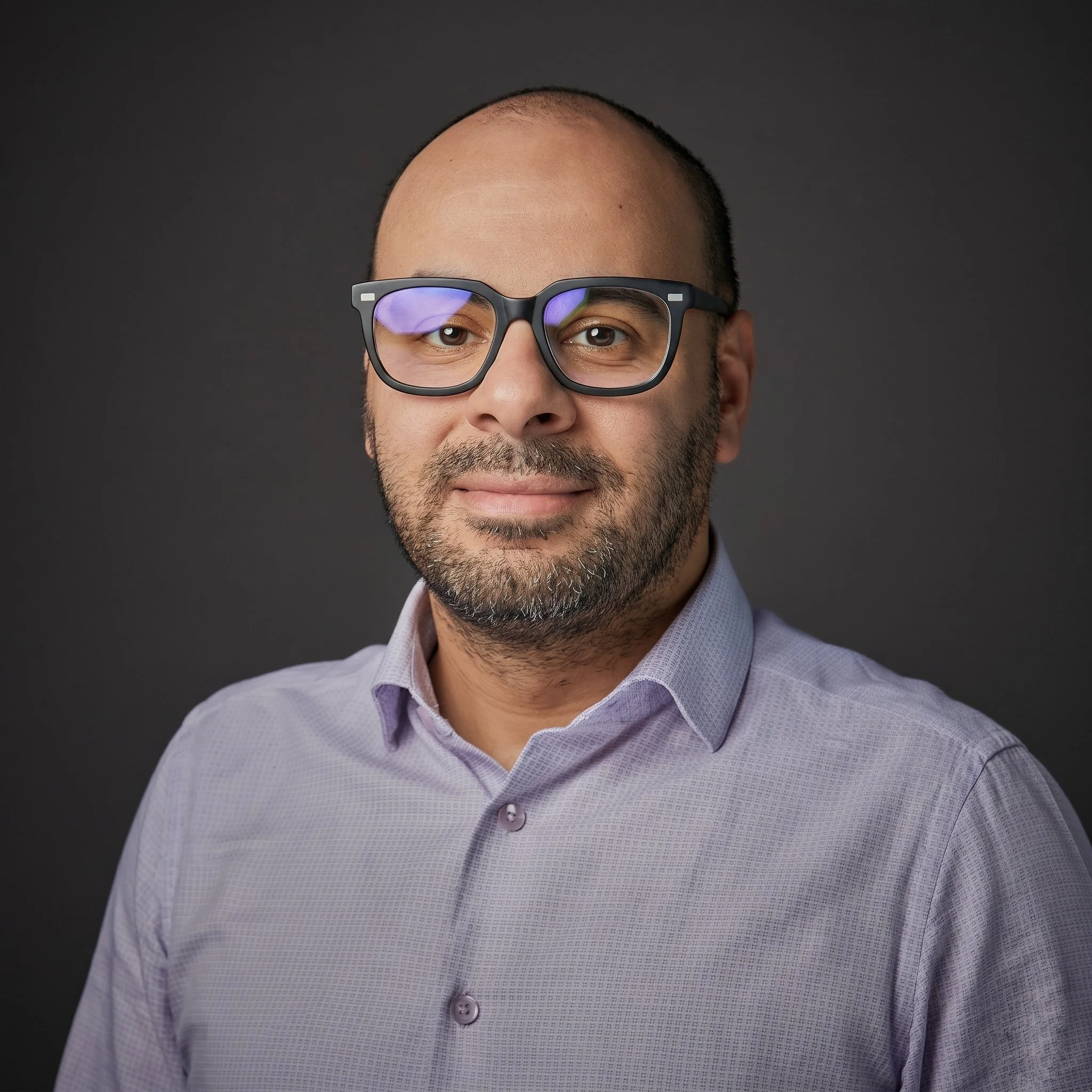 Close-up of a smiling man with short hair, glasses, and a light purple button-up shirt, against a dark background.