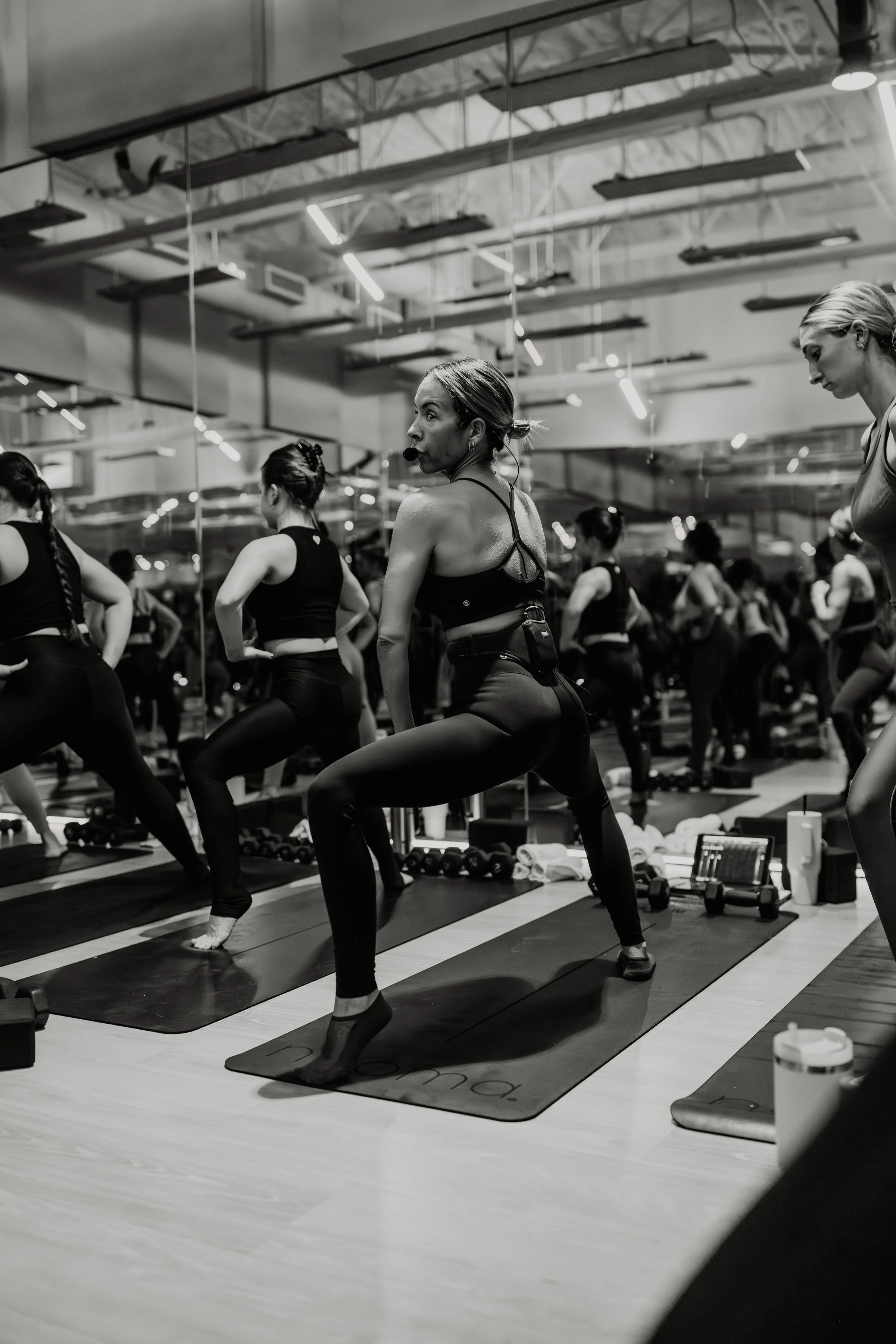 Women attending a fitness or yoga class in a gym, practicing squats on exercise mats with mirrors reflecting their movements.