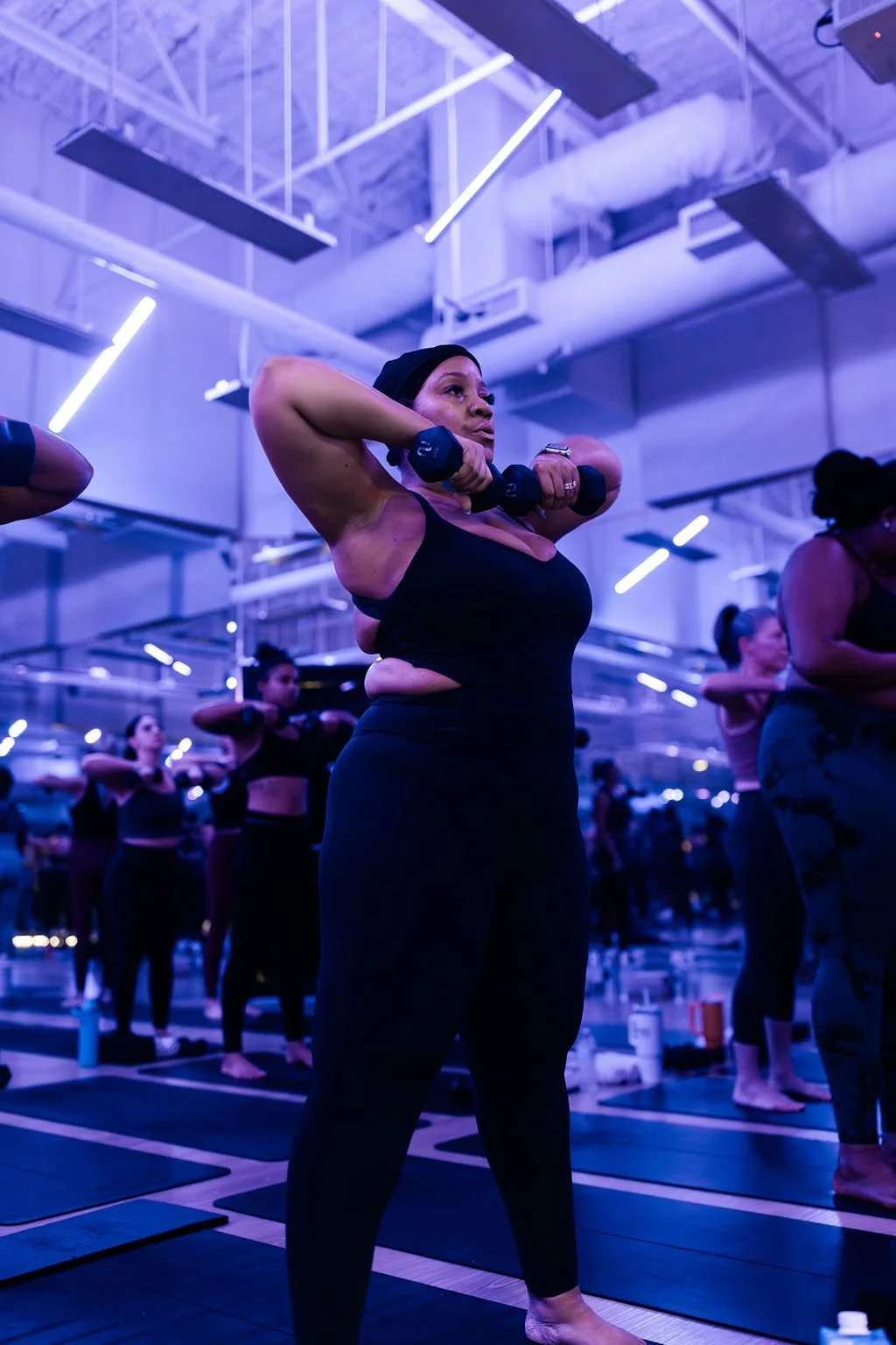 A woman doing a yoga pose with dumbbells in a fitness class, surrounded by other participants, in a dimly lit studio with purple lighting and ceiling lights.