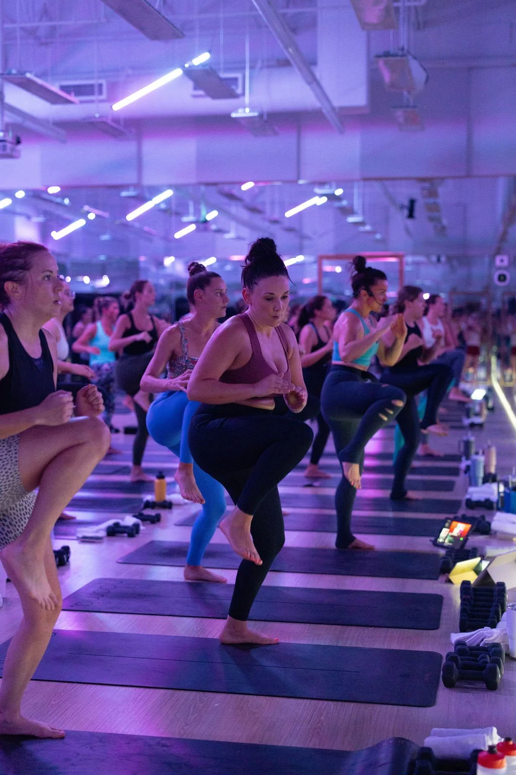 Women participating in a yoga class in a dimly lit studio with purple lighting. They are standing on black yoga mats, lifting one knee and holding their hands together in front of their chest.