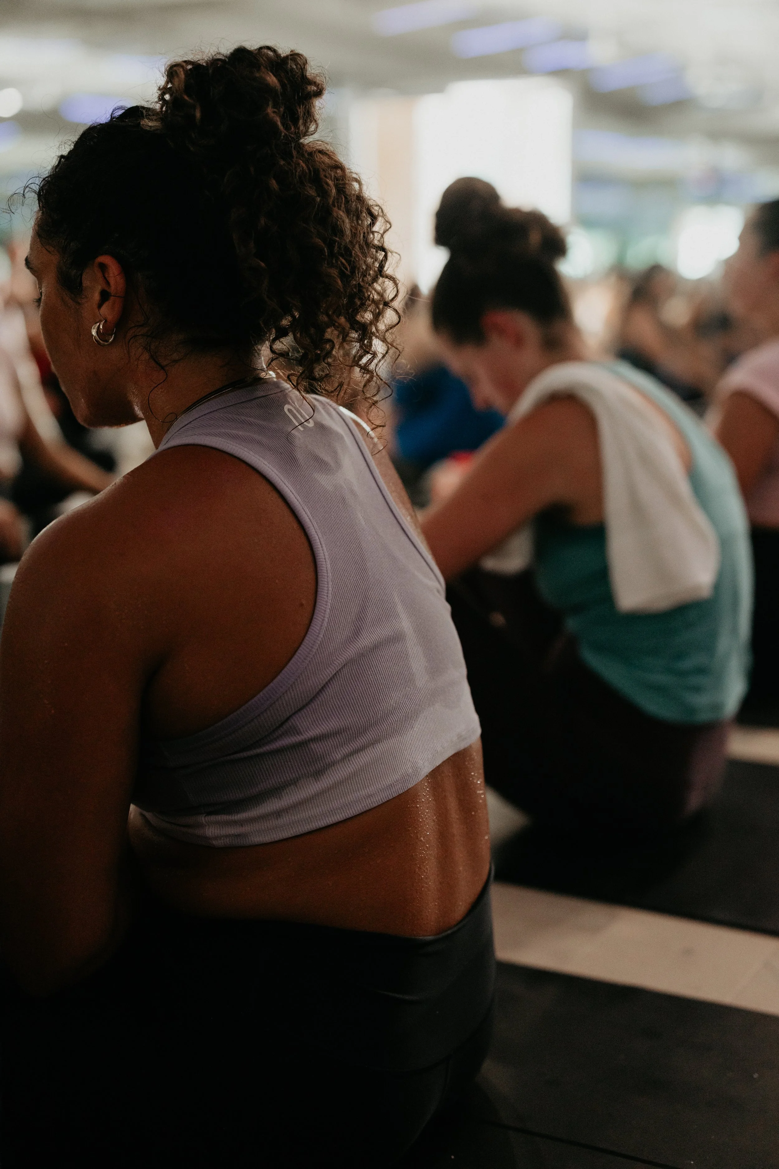 Close-up of a woman with curly hair tied up in a bun, wearing a workout top, in a fitness class or gym setting with others in the background.