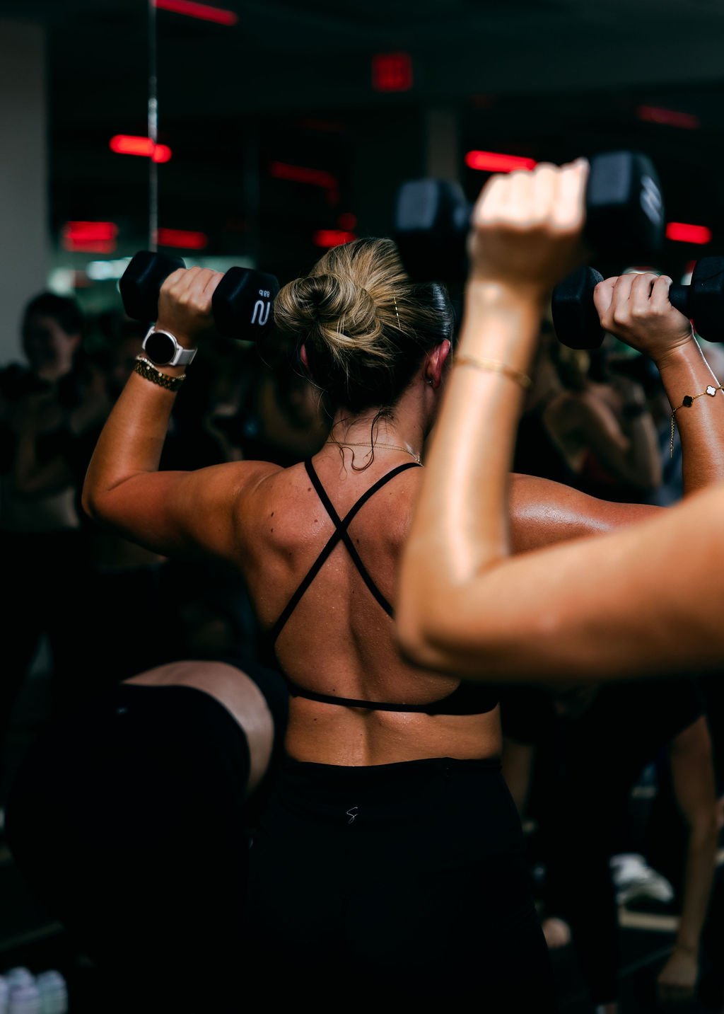 A woman with a bun hairstyle lifting dumbbells during a workout in a gym, with other people in the background.