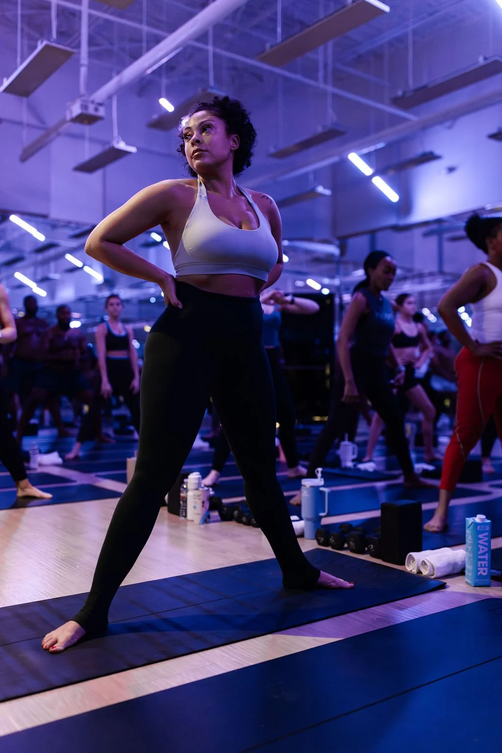 A woman participating in a yoga class, standing on a yoga mat with her hands on her hips, surrounded by other women in a dimly lit studio with purple lighting.