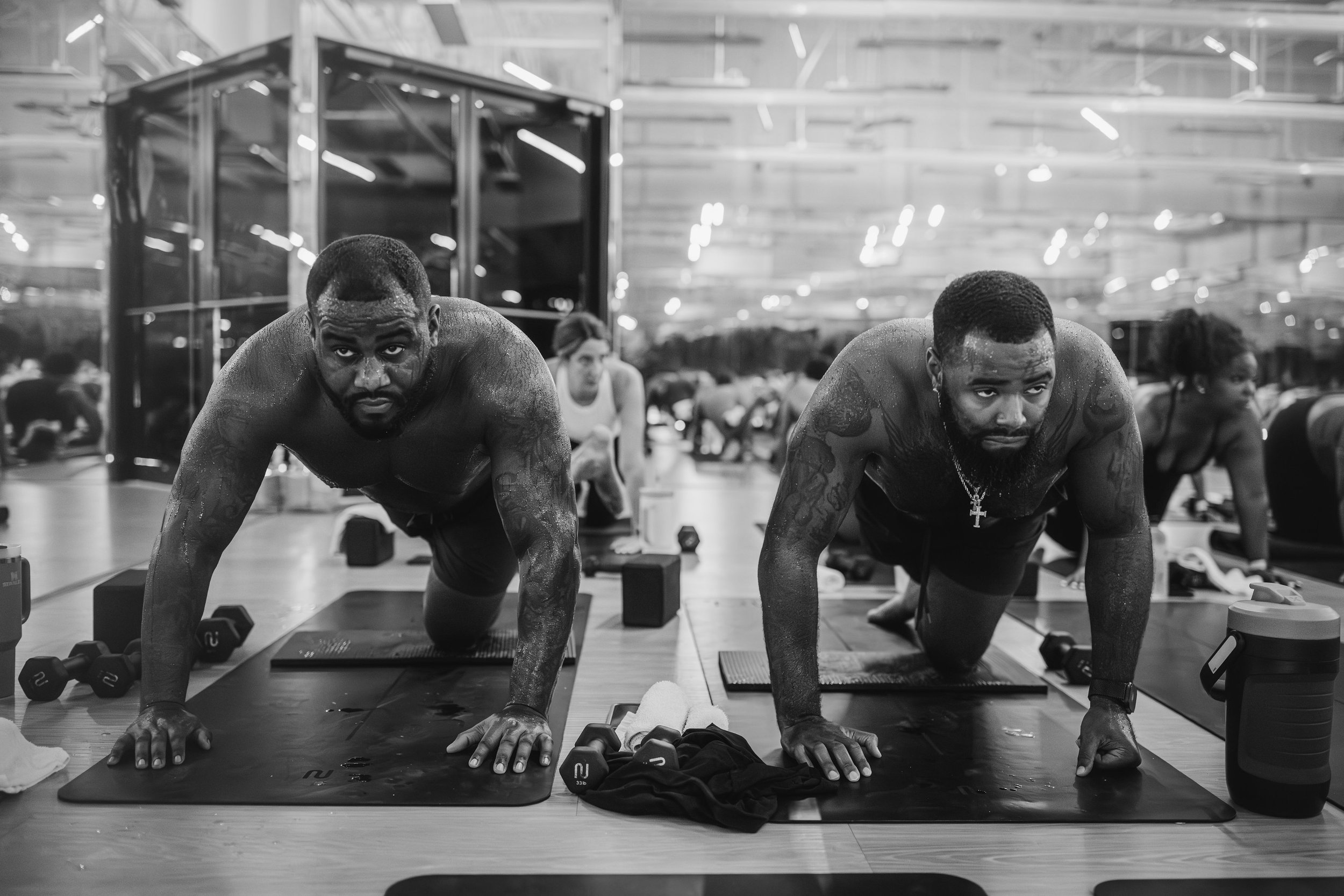 Two Black men performing push-ups on mats in a gym, with weights nearby, surrounded by others exercising in the background, in black and white.