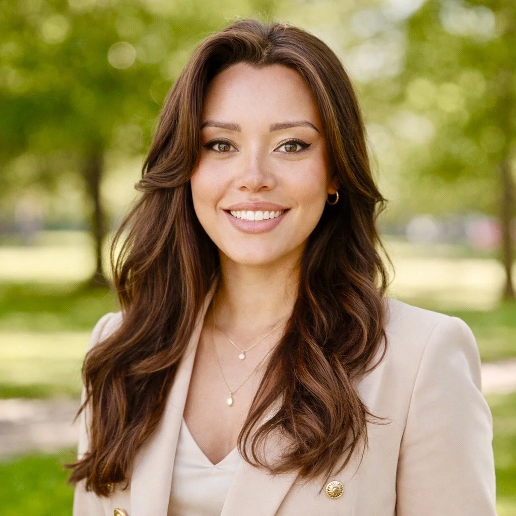 A young woman with long, wavy brown hair smiling outdoors in a park with green trees in the background.
