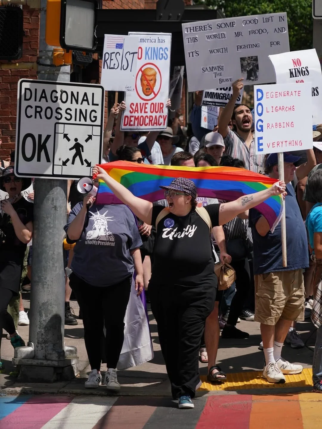 A crowd of protesters at a rally holding signs and a rainbow flag, advocating for LGBTQ rights and criticizing political figures, with a woman at the front waving the rainbow flag and smiling.