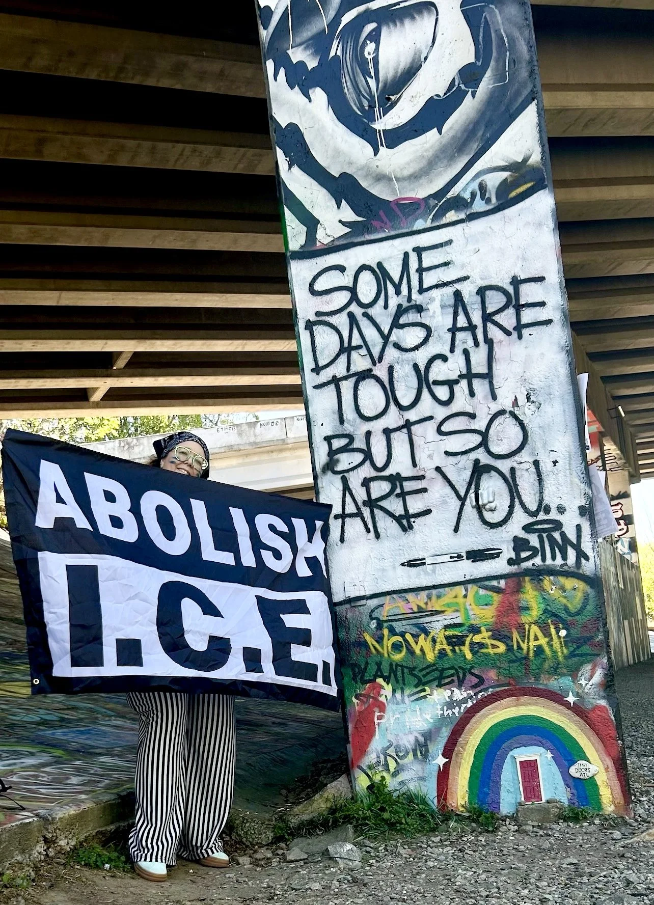 A person holding a large protest sign reading "ABOUTISE" with colors black and white, standing under an overpass with graffiti and posters on a concrete wall. The individual is wearing glasses, a black top, striped pants, and has a headscarf.