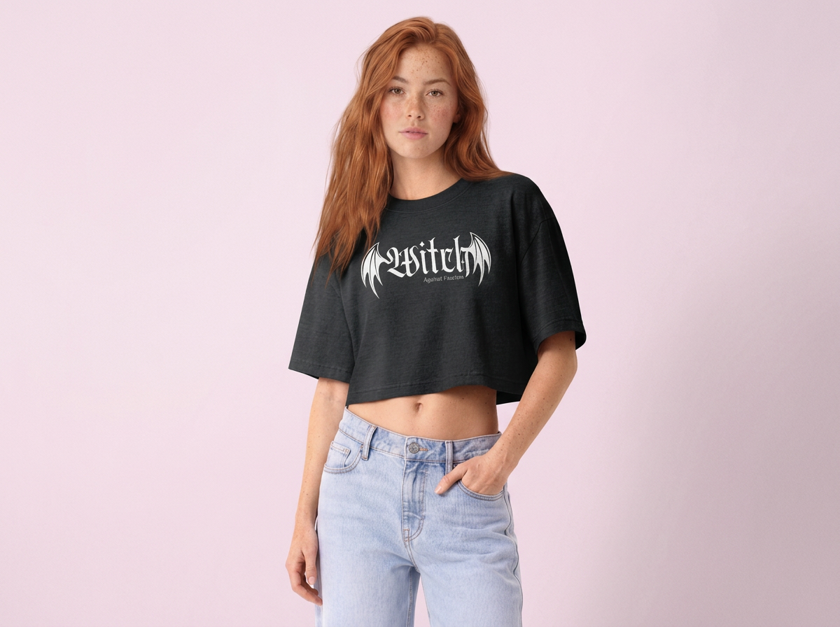 A young woman with long red hair, wearing a black crop top with white Gothic-style text and wing design, and light blue high-waisted jeans, standing against a light pink background.