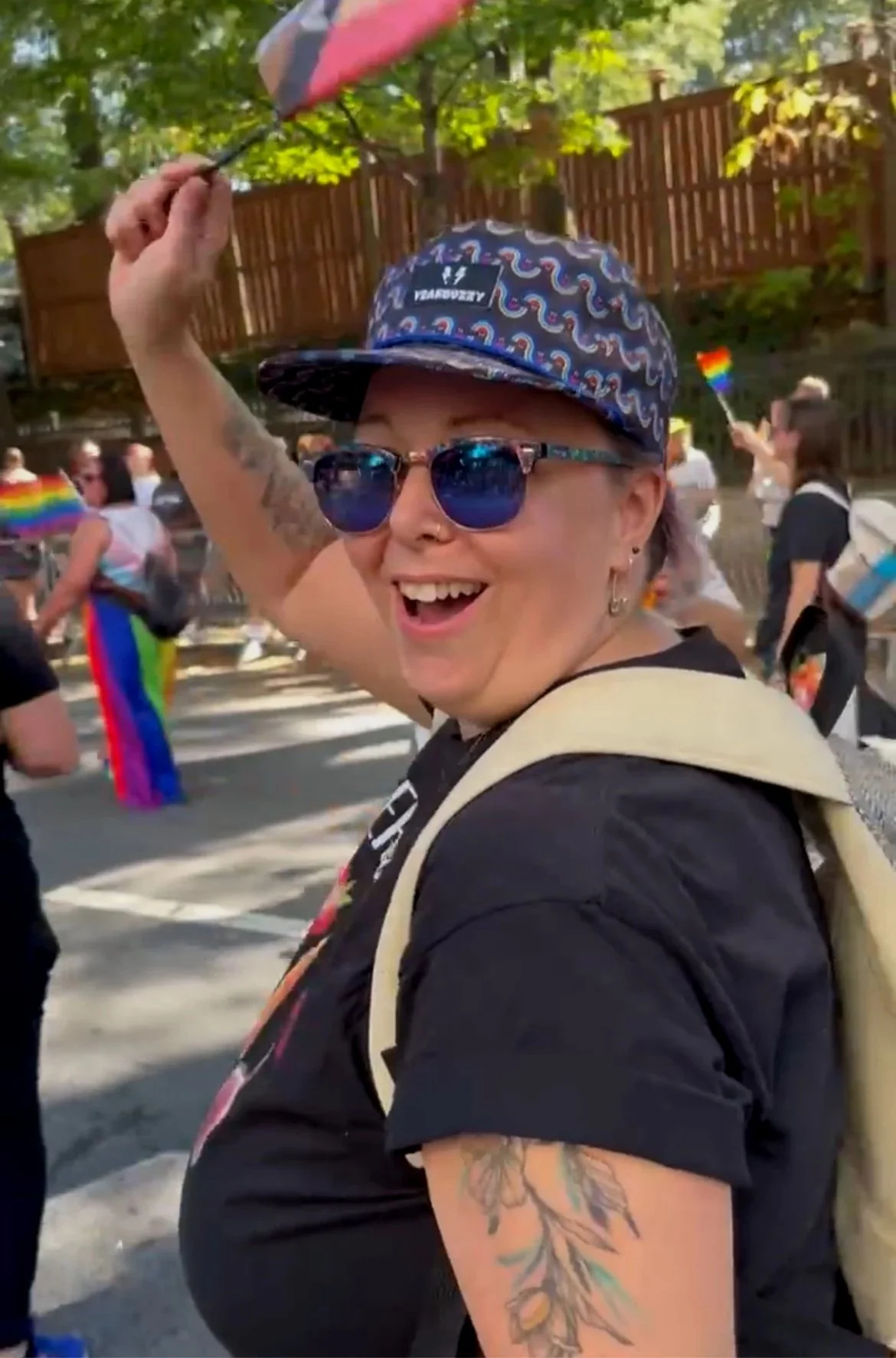A smiling person with tattoos on their arm, wearing sunglasses and a patterned cap, holding an umbrella, participating in an outdoor pride parade.