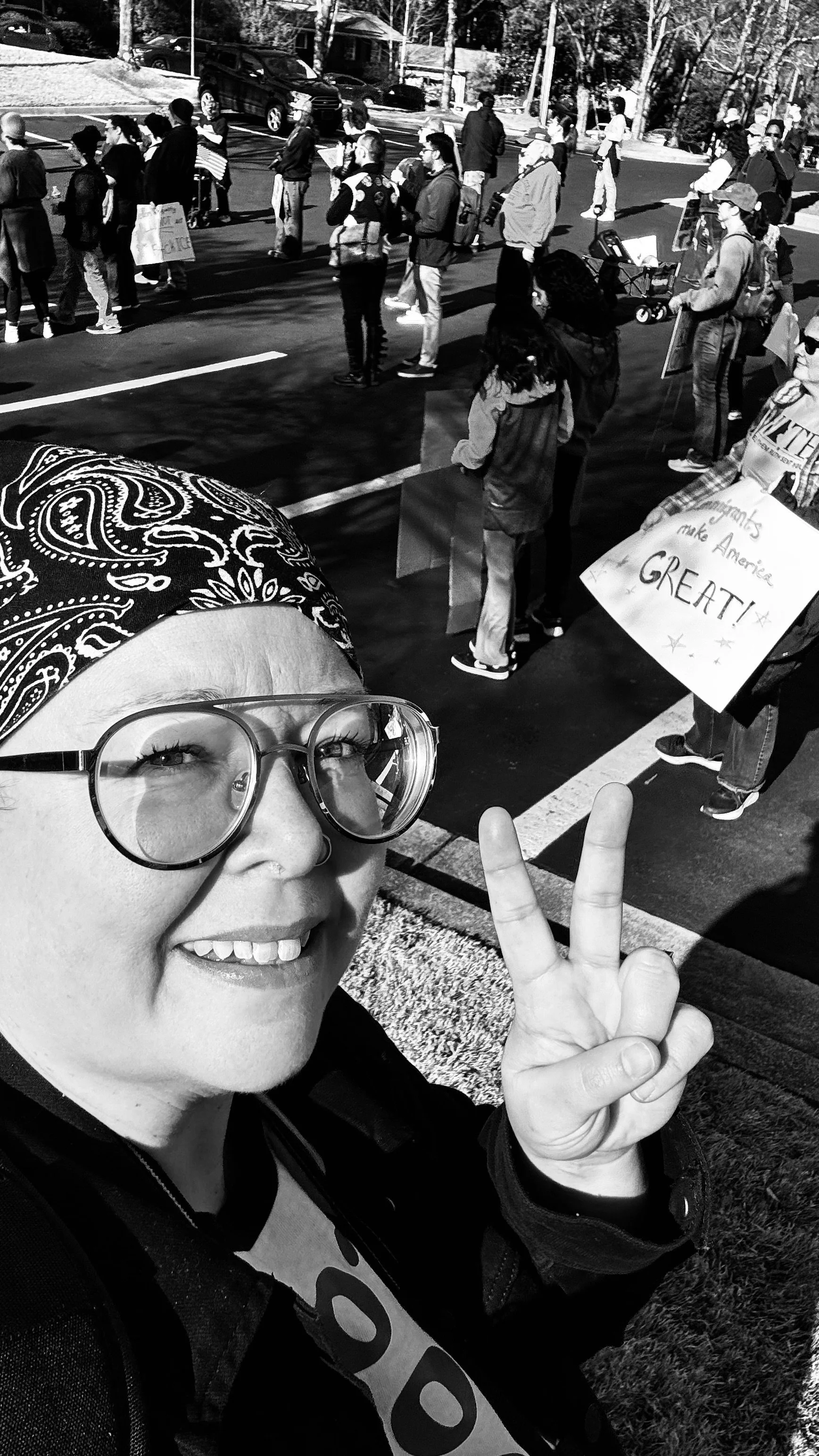 A woman taking a selfie at a protest or rally, making a peace sign with her hand. The crowd in the background holds signs, including one that says "Humans Make America Great!"