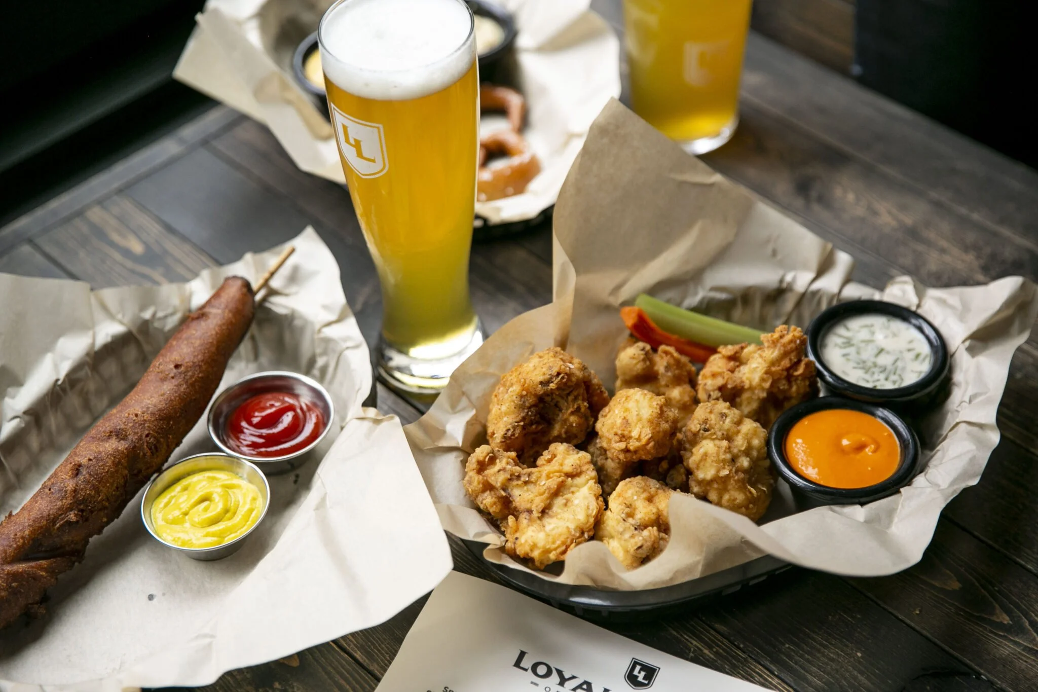 A spread of fried chicken pieces, a cheeseburger with toppings, fried mozzarella sticks, two glasses of beer, and various dipping sauces on a wooden table.