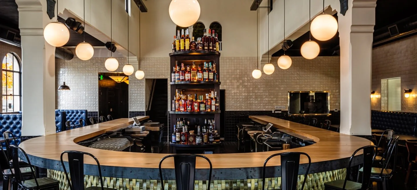 Interior of a modern bar with a curved wooden bar counter, black chairs, hanging globe lights, a central shelf with bottles of alcohol, and a mix of white and black tiled walls.