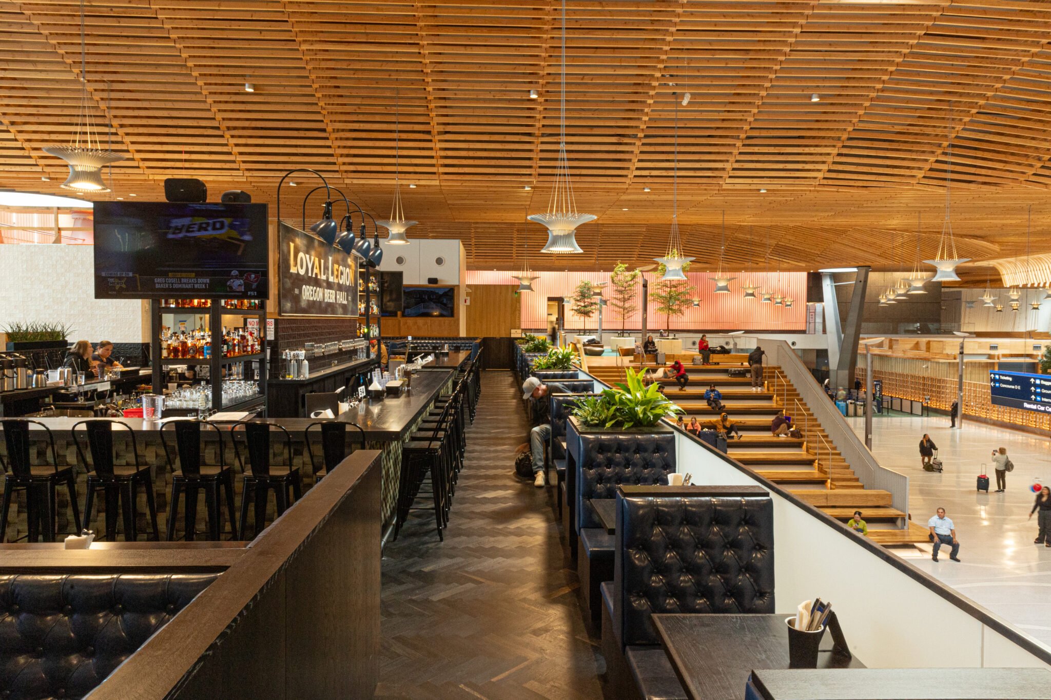 Interior of a modern airport with wooden ceiling, bar area, seating, and people walking and sitting.