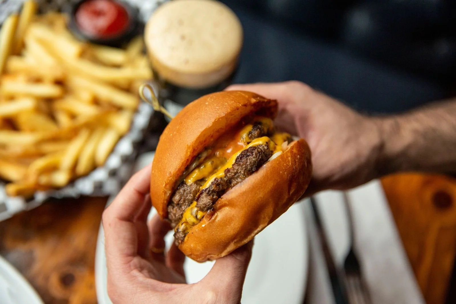 Person holding a cheeseburger with a side of French fries and condiments in the background.