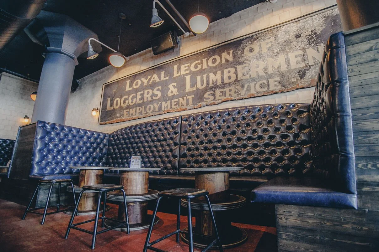 Vintage-style bar or restaurant interior with a large distressed sign reading 'Loyal Legion of Loggers & Lumbermen Employment Service,' dark tufted leather booth seating around wooden tables, and industrial lighting fixtures.