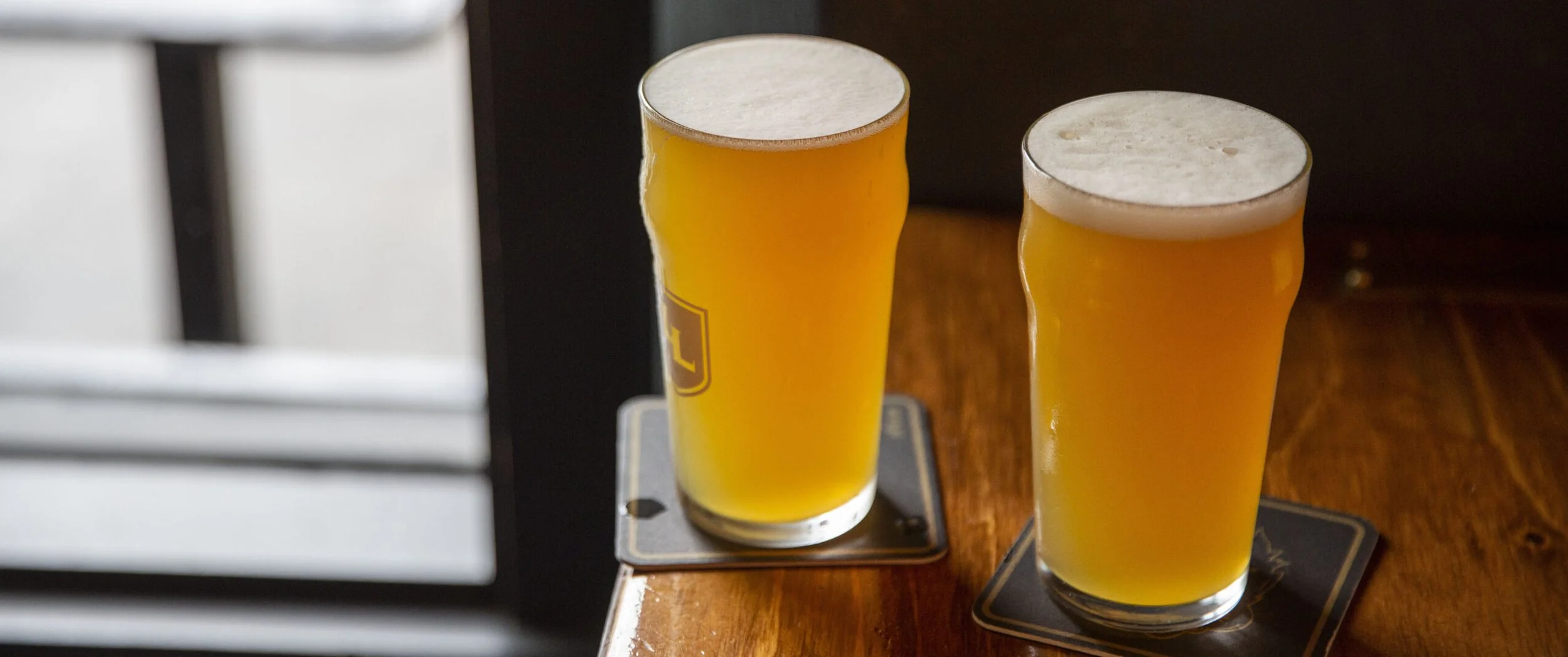 Two tall glasses of pale yellow beer with foamy heads on wooden bar counter, blurred background.