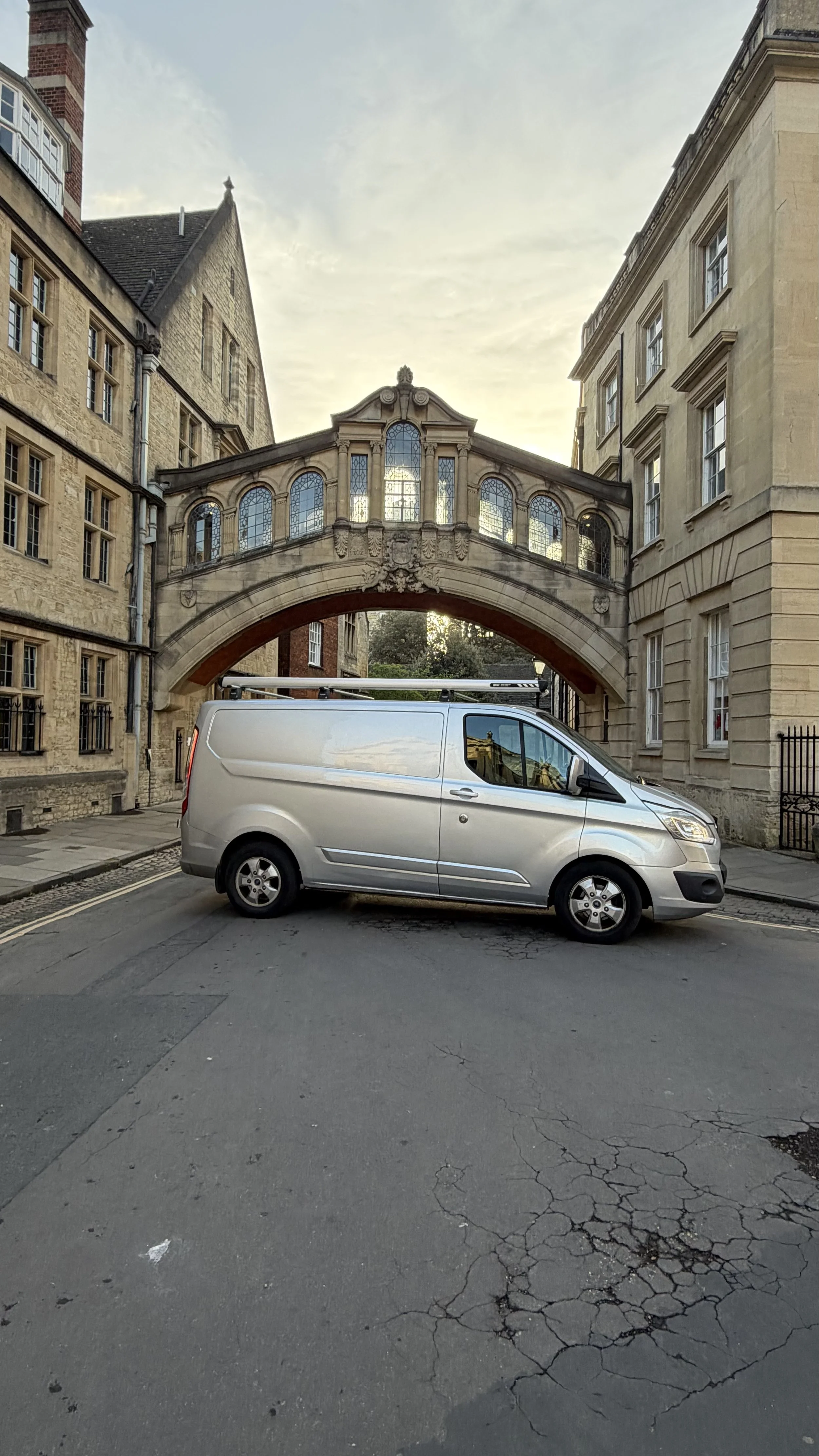 A silver van parked on a street beneath a historic stone bridge connecting two ornate buildings, with cracked pavement in the foreground and a partly cloudy sky above.