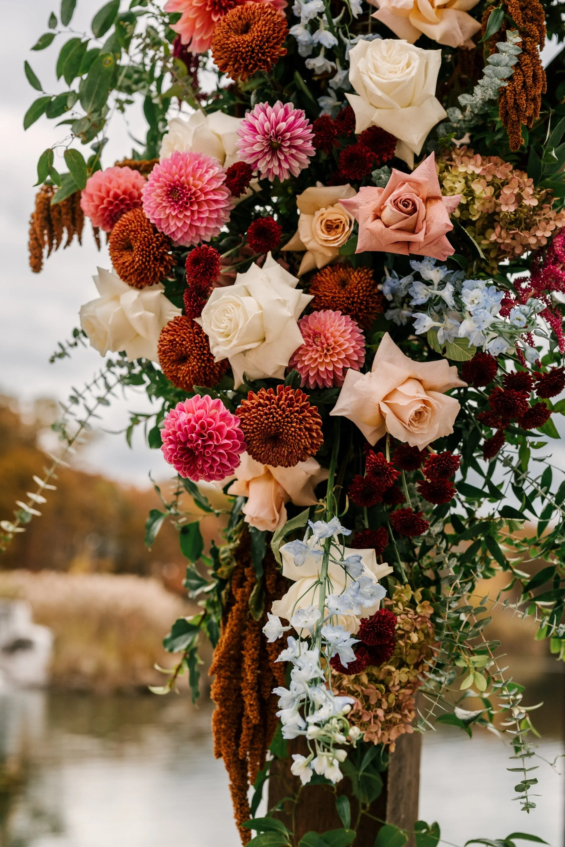 A colorful floral arrangement with pink dahlias, white roses, red and orange flowers, blue accents, and green foliage, set outdoors near water with a blurred background.
