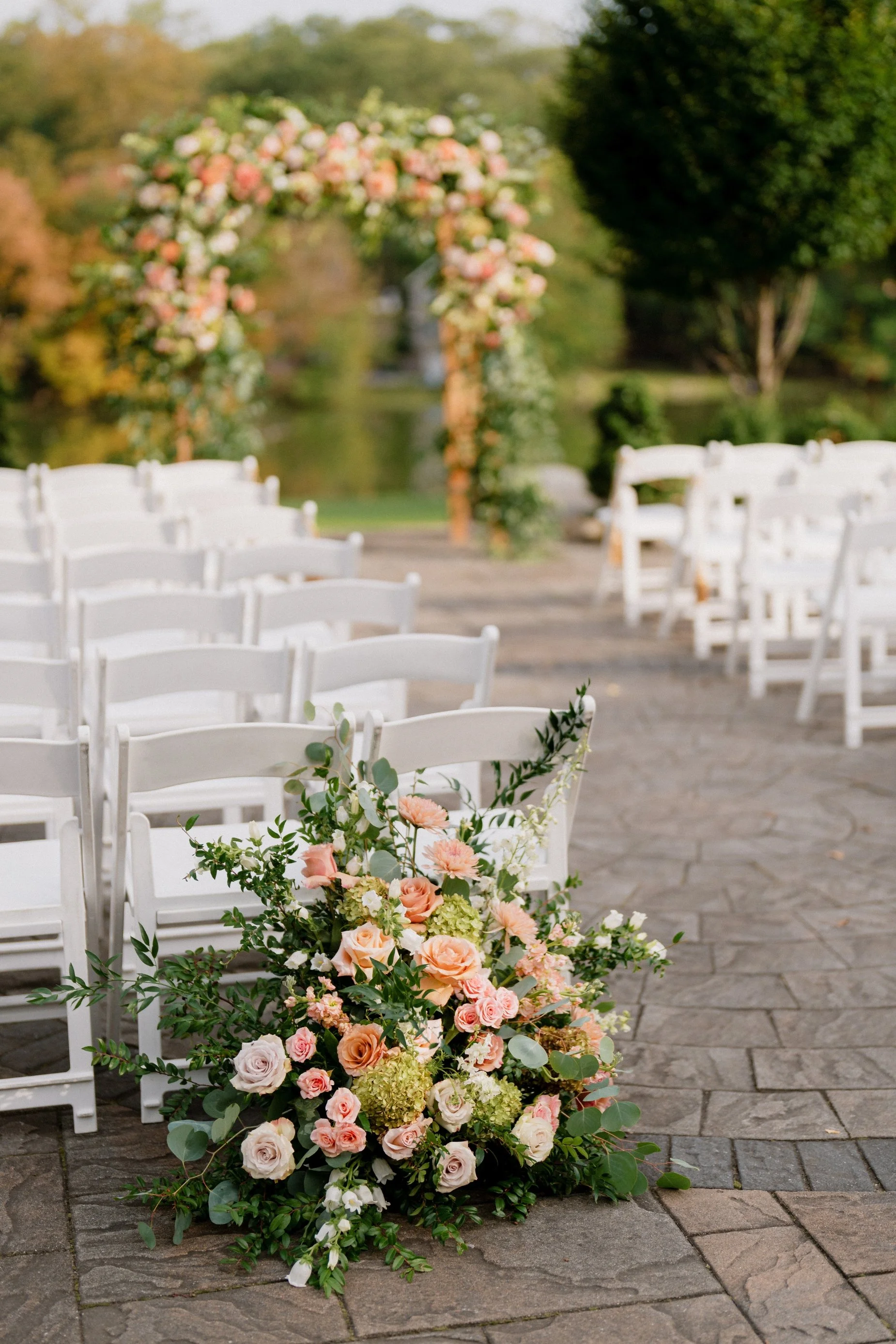 White chairs arranged outdoors on a brick patio with a floral arrangement featuring pink, peach, and white roses and greenery at the front. An arch decorated with similar flowers can be seen in the background, with trees and a lake or pond nearby.