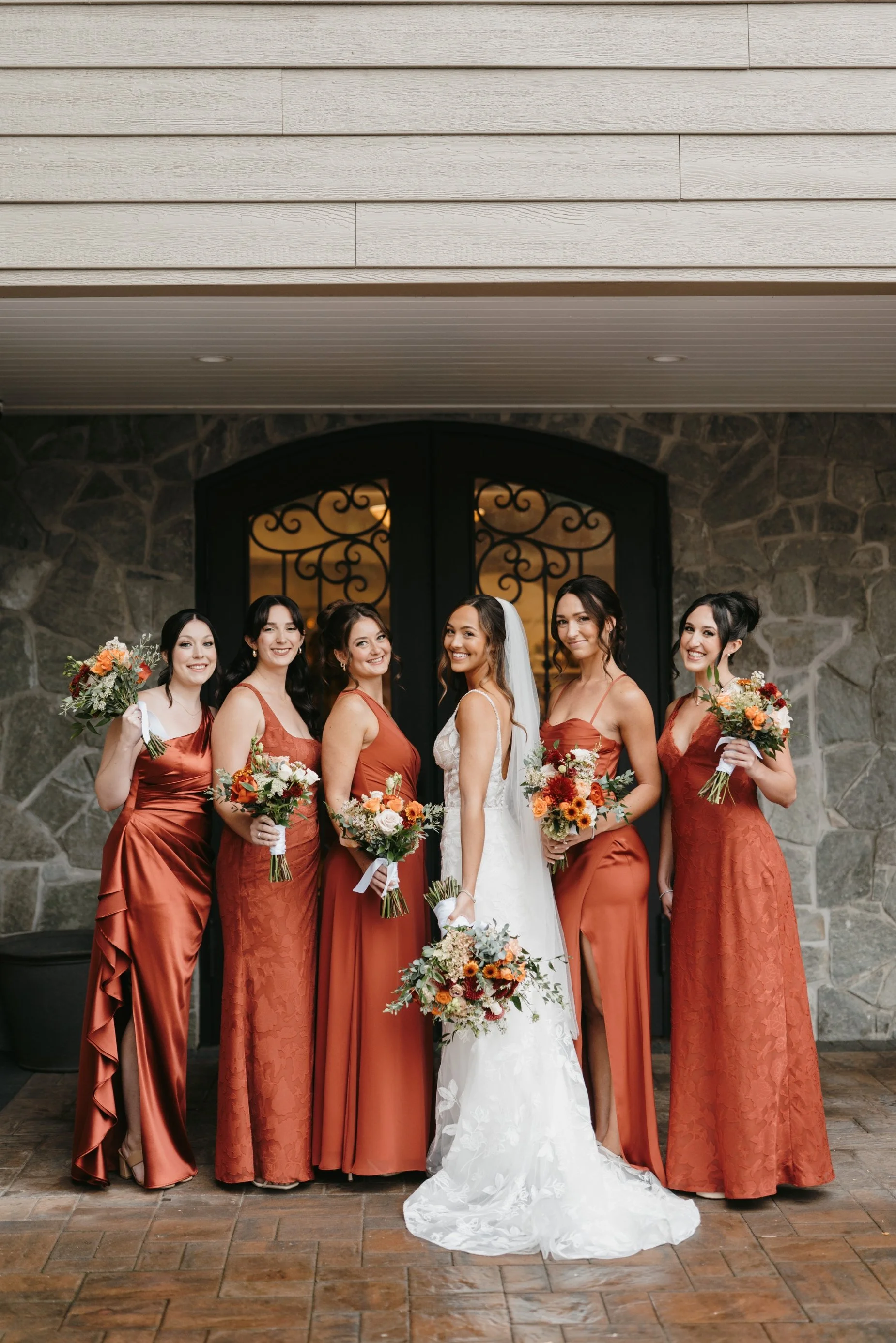 A bride in a white wedding dress standing with six women dressed in matching rust-colored bridesmaid dresses, holding bouquets, standing in front of a large stone and wood door with wrought iron details.