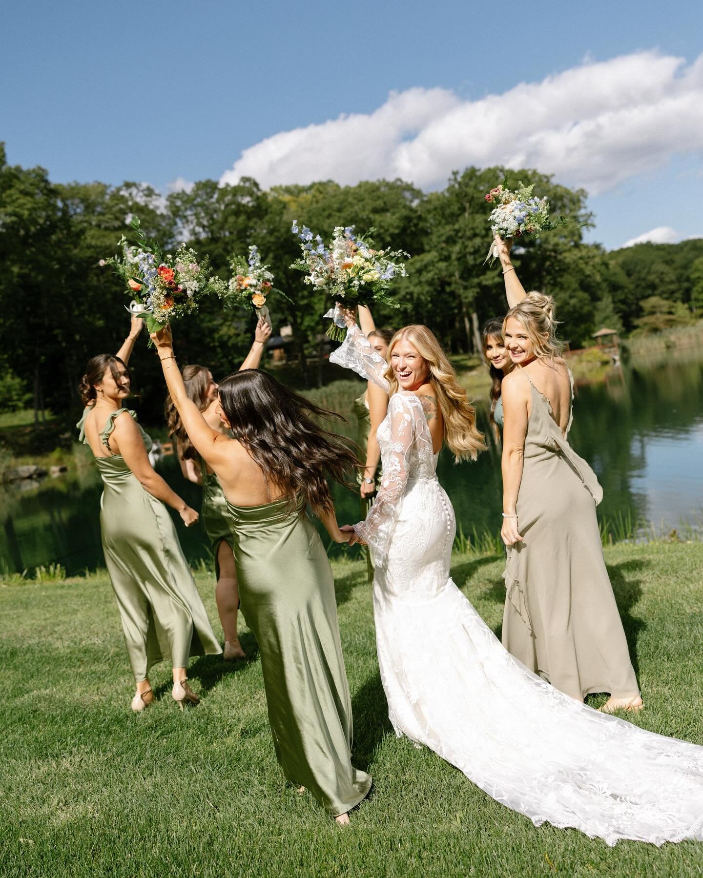 Group of women celebrating outdoors by a lake, holding bouquets of flowers, with trees and blue sky in the background.
