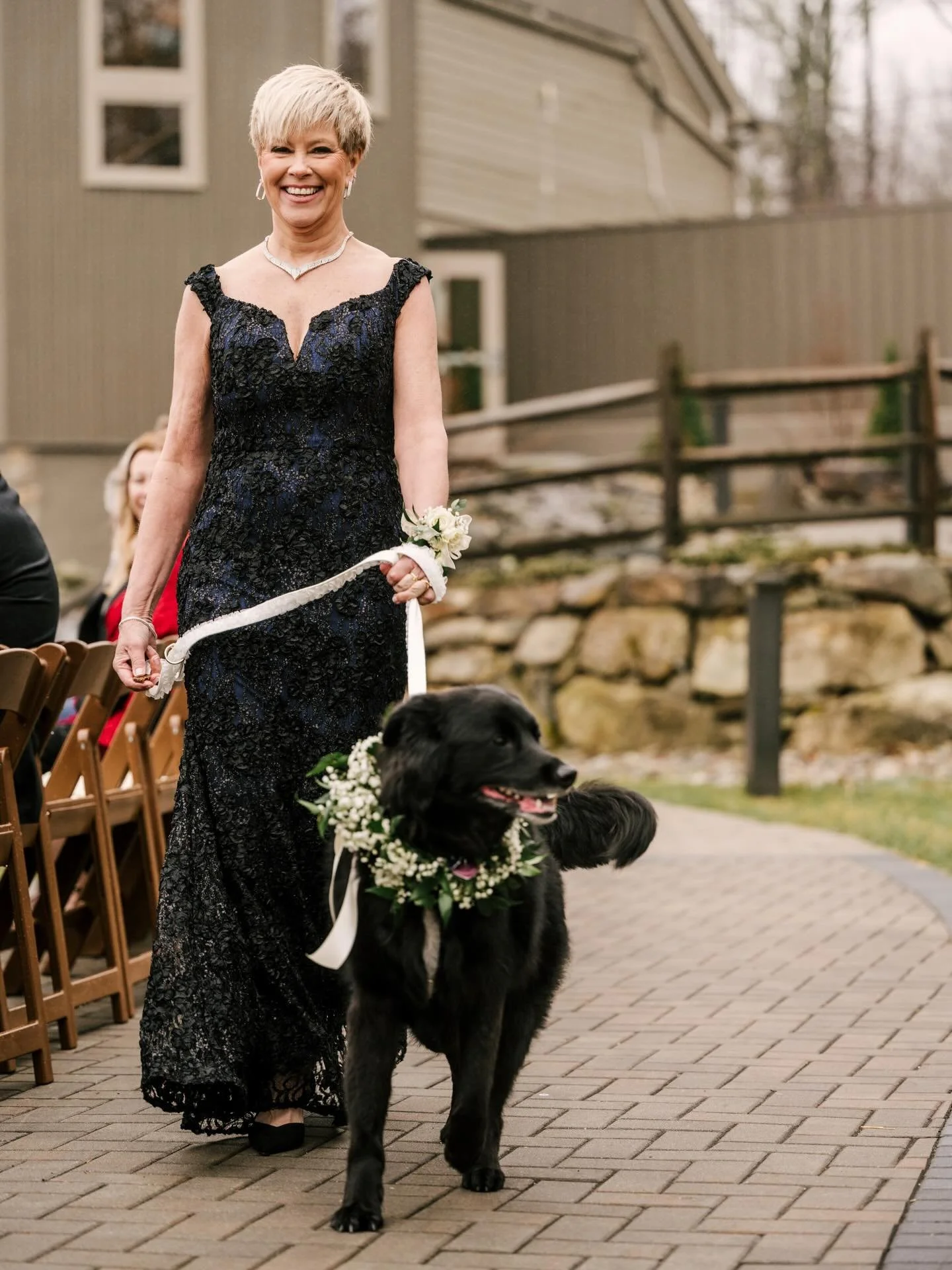 A woman in a black lace dress smiling and holding a leash attached to a black dog decorated with a floral wreath walking outdoors on a brick path, with a house and a fence in the background.