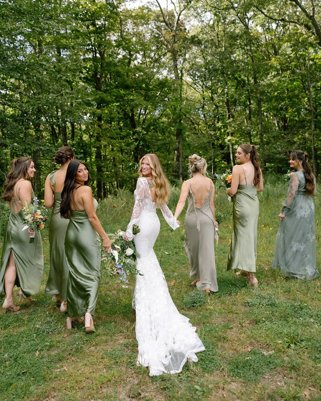 A bride in a white lace wedding dress holding a bouquet, walking in a line with six bridesmaids in pastel green, gray, and blue dresses in a forested outdoor setting.