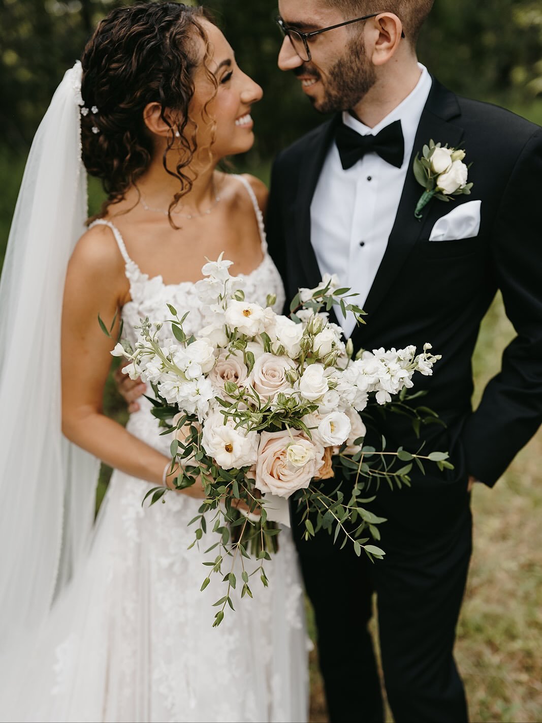 A bride and groom smiling and looking at each other outdoors, the bride holding a large bouquet of white and blush roses and greenery, both dressed in wedding attire.