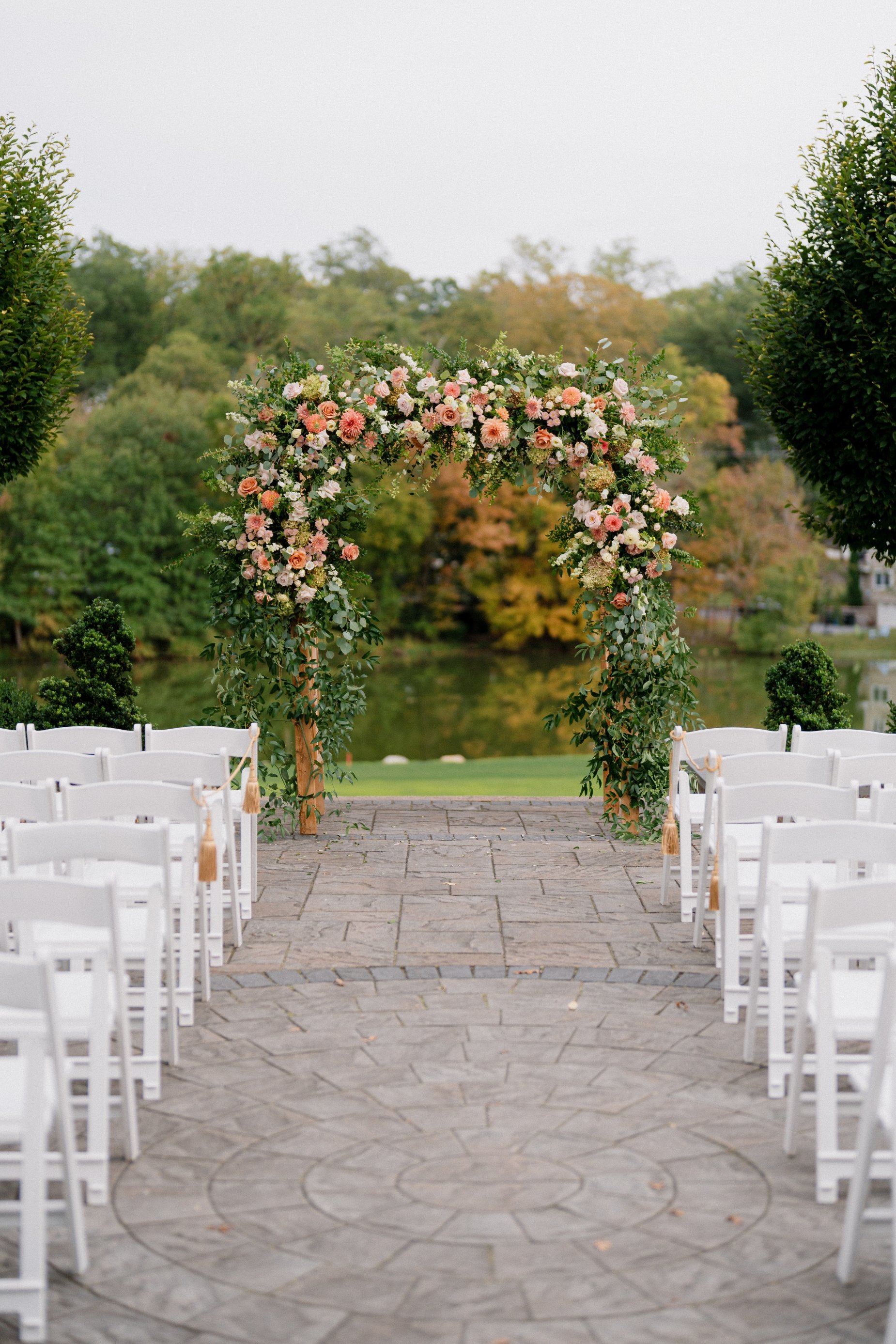 Outdoor wedding ceremony setup with white chairs arranged on either side of a stone aisle, a floral arch with pink, peach, and white flowers at the end of the aisle, and green trees in the background.