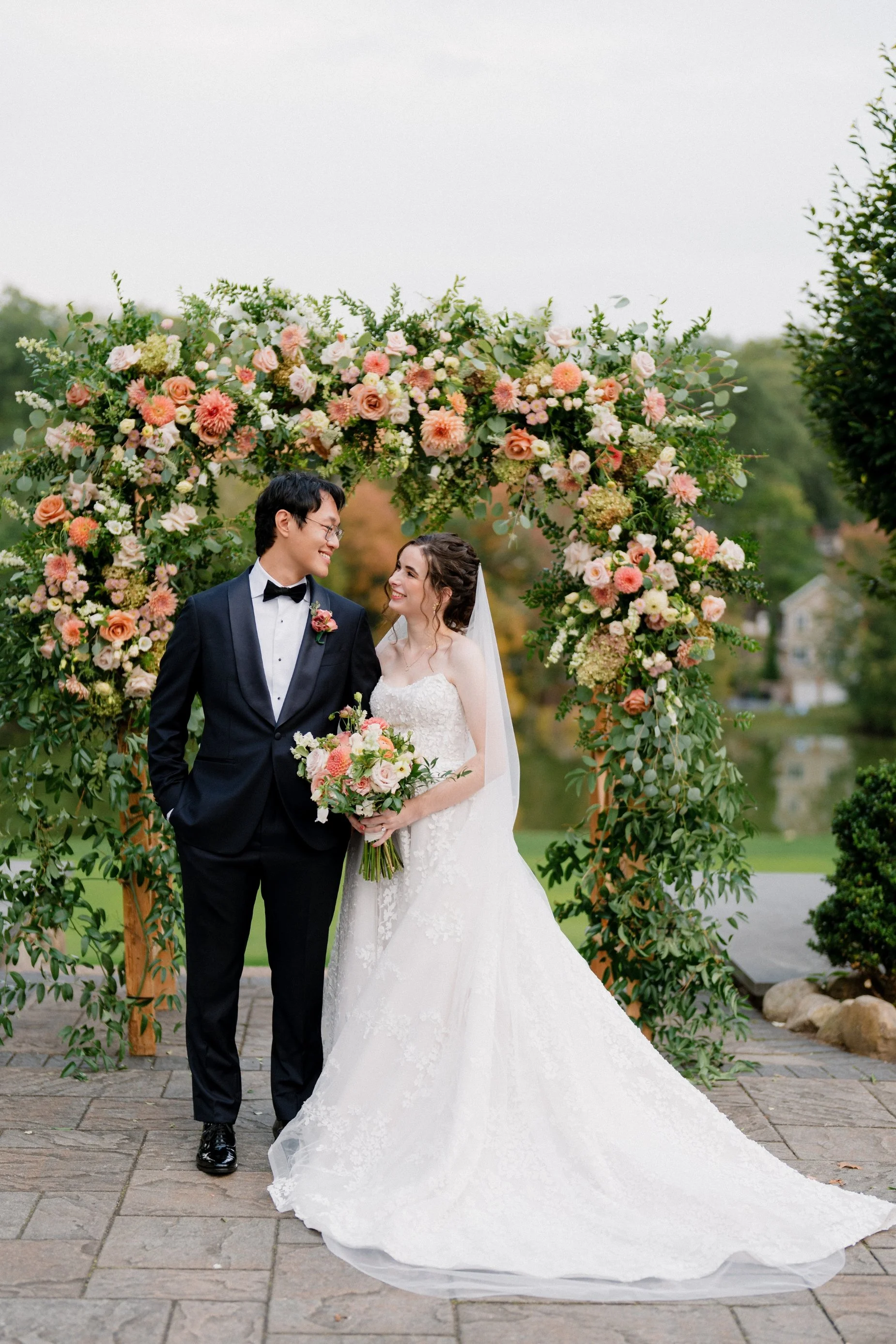 A bride and groom smiling at each other under a large flower arch on a paved outdoor setting during their wedding.