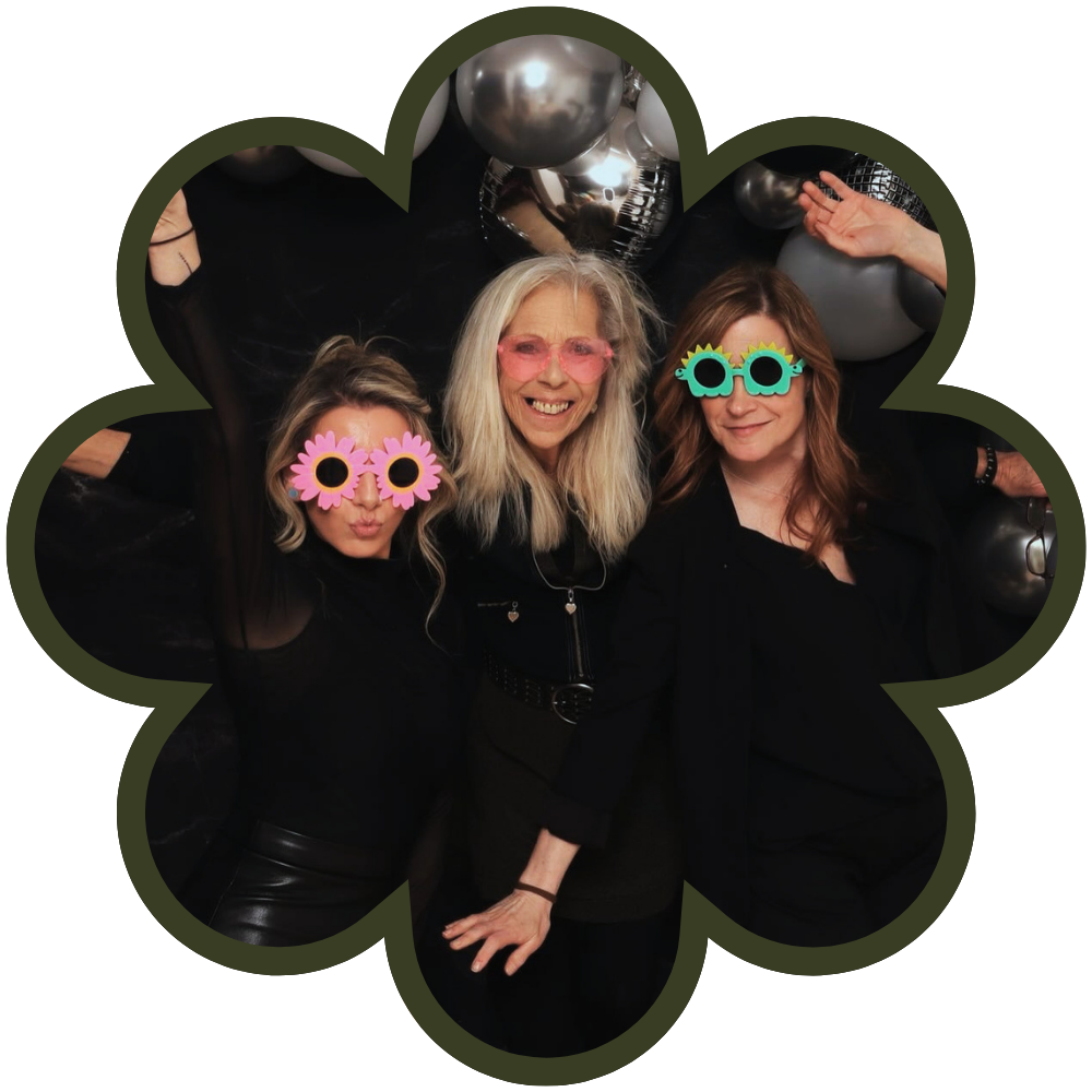 Three women wearing colorful sunglasses and black clothing, smiling and posing at a party with metallic balloons in the background.