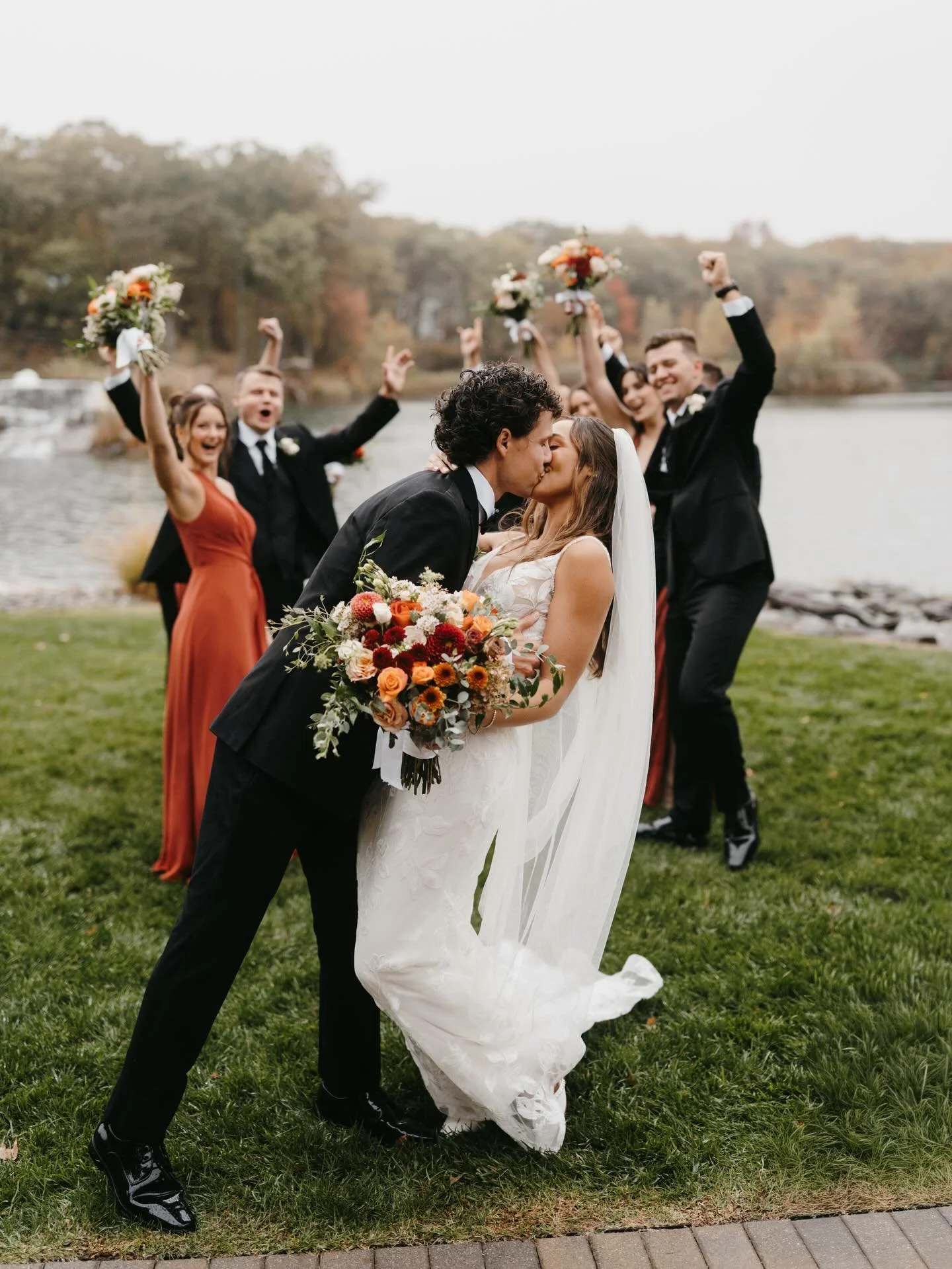 A bride and groom kissing at their outdoor wedding, surrounded by joyful bridesmaids and groomsmen celebrating