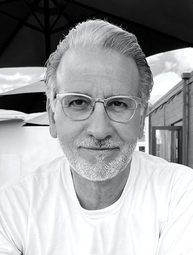 A black and white photo of a smiling man with glasses, a beard, and gray hair, sitting outdoors under a canopy with a building and umbrella in the background.