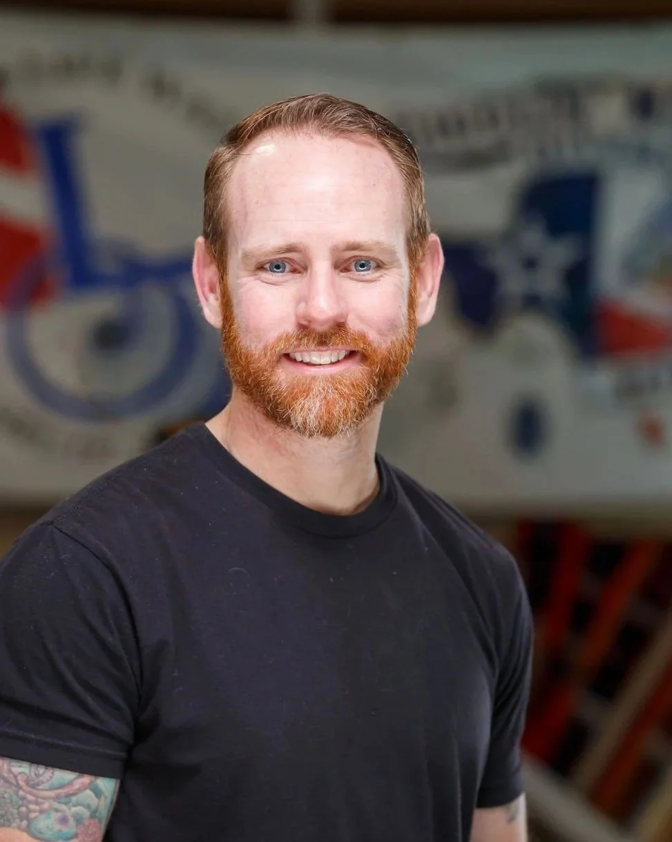 A smiling man with red hair and beard, wearing a black T-shirt, standing in front of a blurred background featuring a logo with an eagle and American flags.