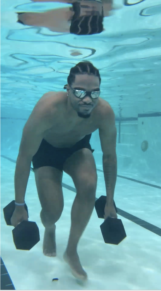 A man swimming underwater in a pool, holding Dumbbells in each hand, wearing sunglasses and black swim shorts.