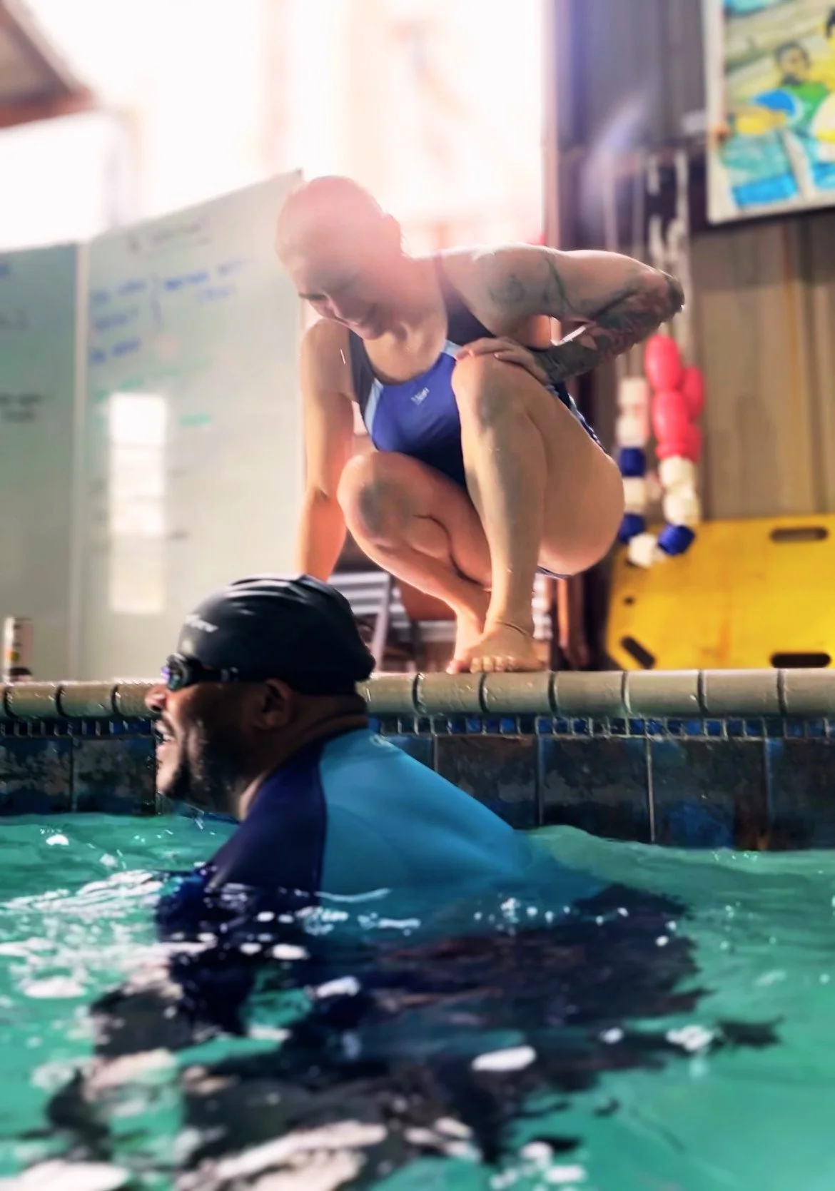 A female swimmer in a swimsuit crouches on the edge of a swimming pool, preparing to dive into the water where a male swimmer in a cap and goggles is swimming.