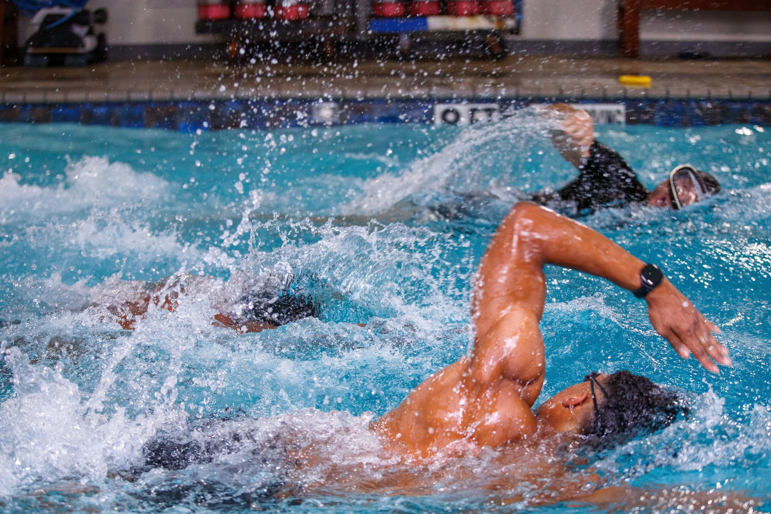 Swimmers competing in a race, with one swimmer in black cap and goggles reaching the water, splashing around in an indoor pool.
