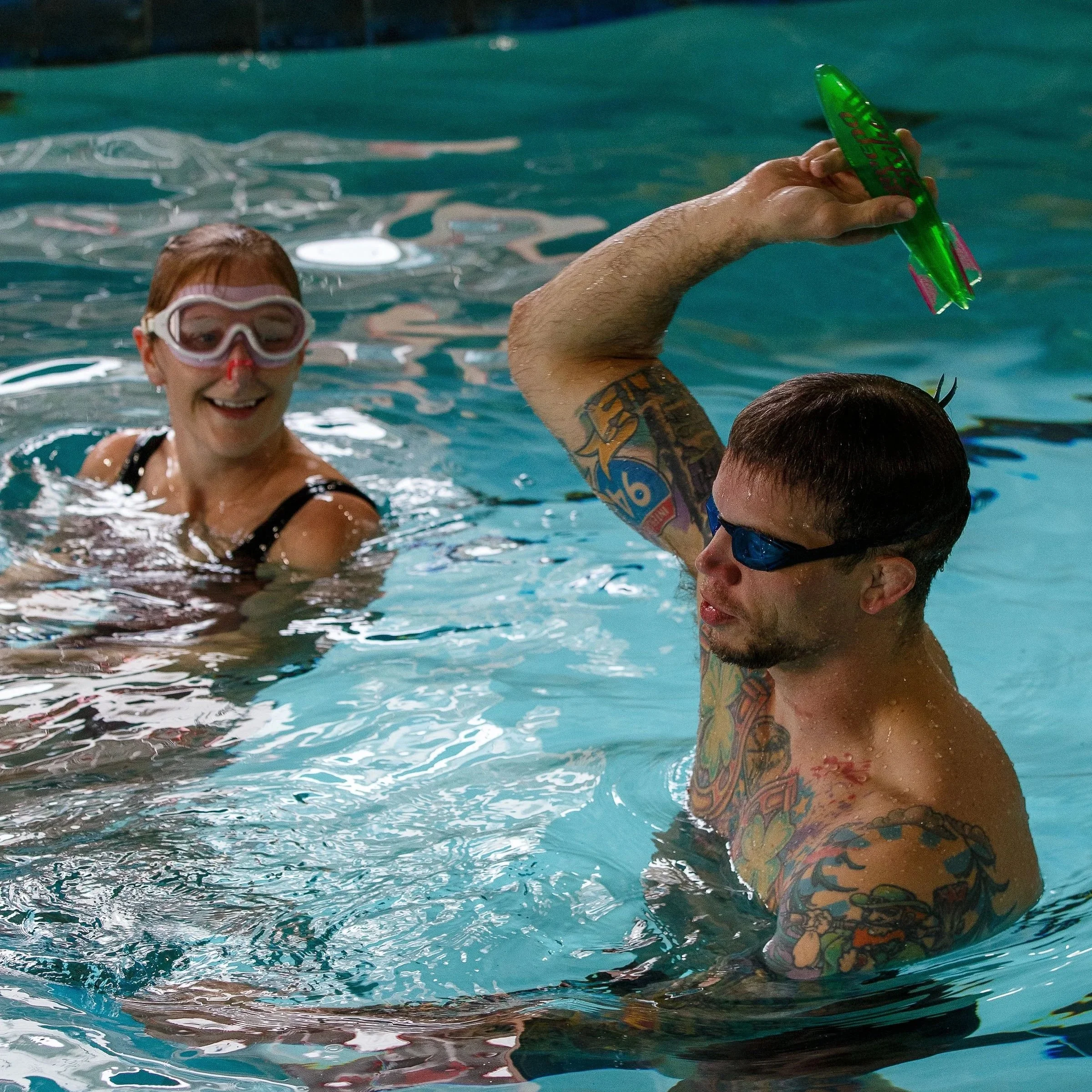 A woman with a dive mask smiling in a swimming pool, holding a green plastic toy, with a man with a tattooed arm.