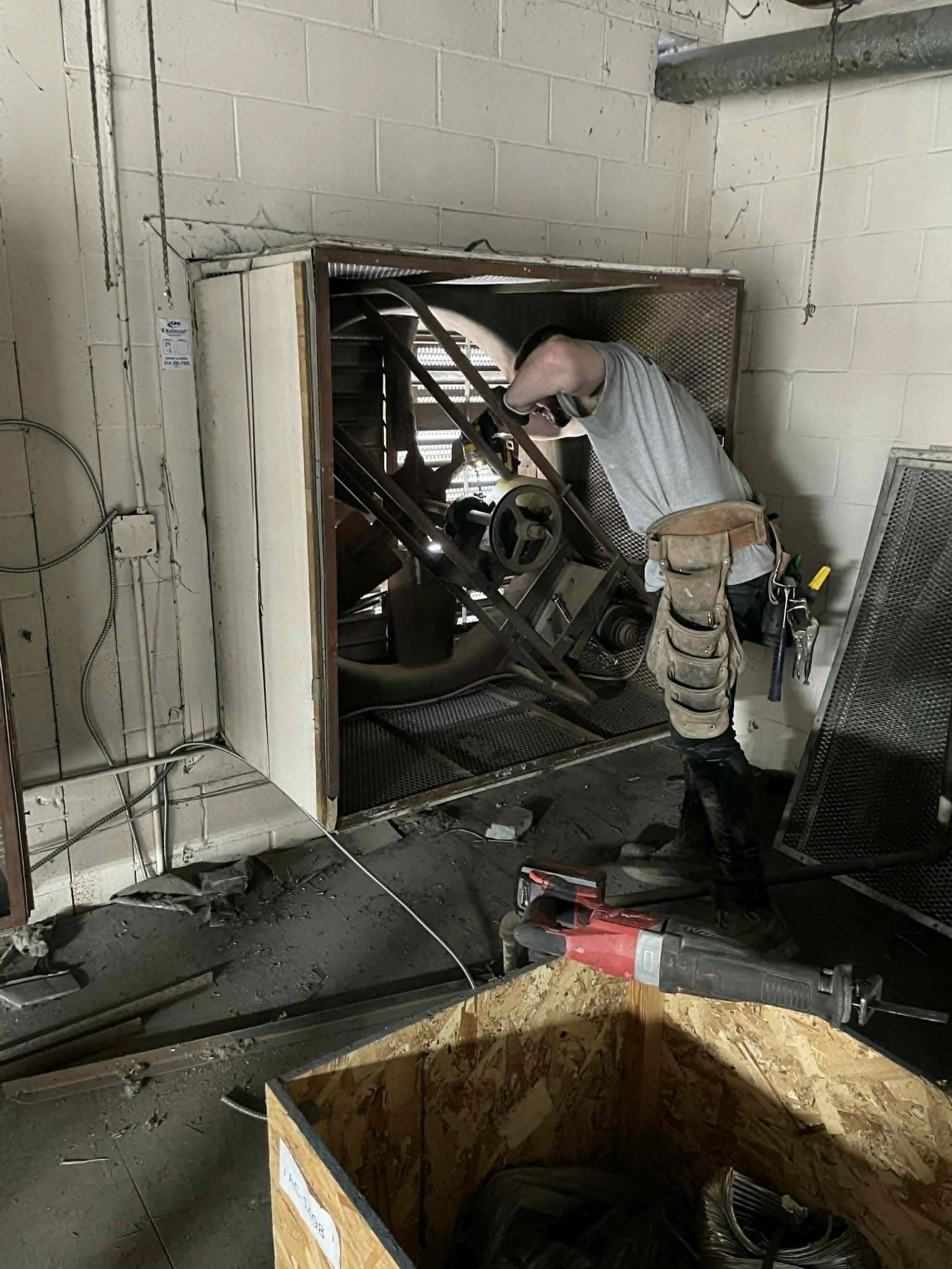 Un homme en train de travailler dans un espace de construction, utilisant des outils électriques, avec un mur en brique en arrière-plan et un sol encombré de déchets.