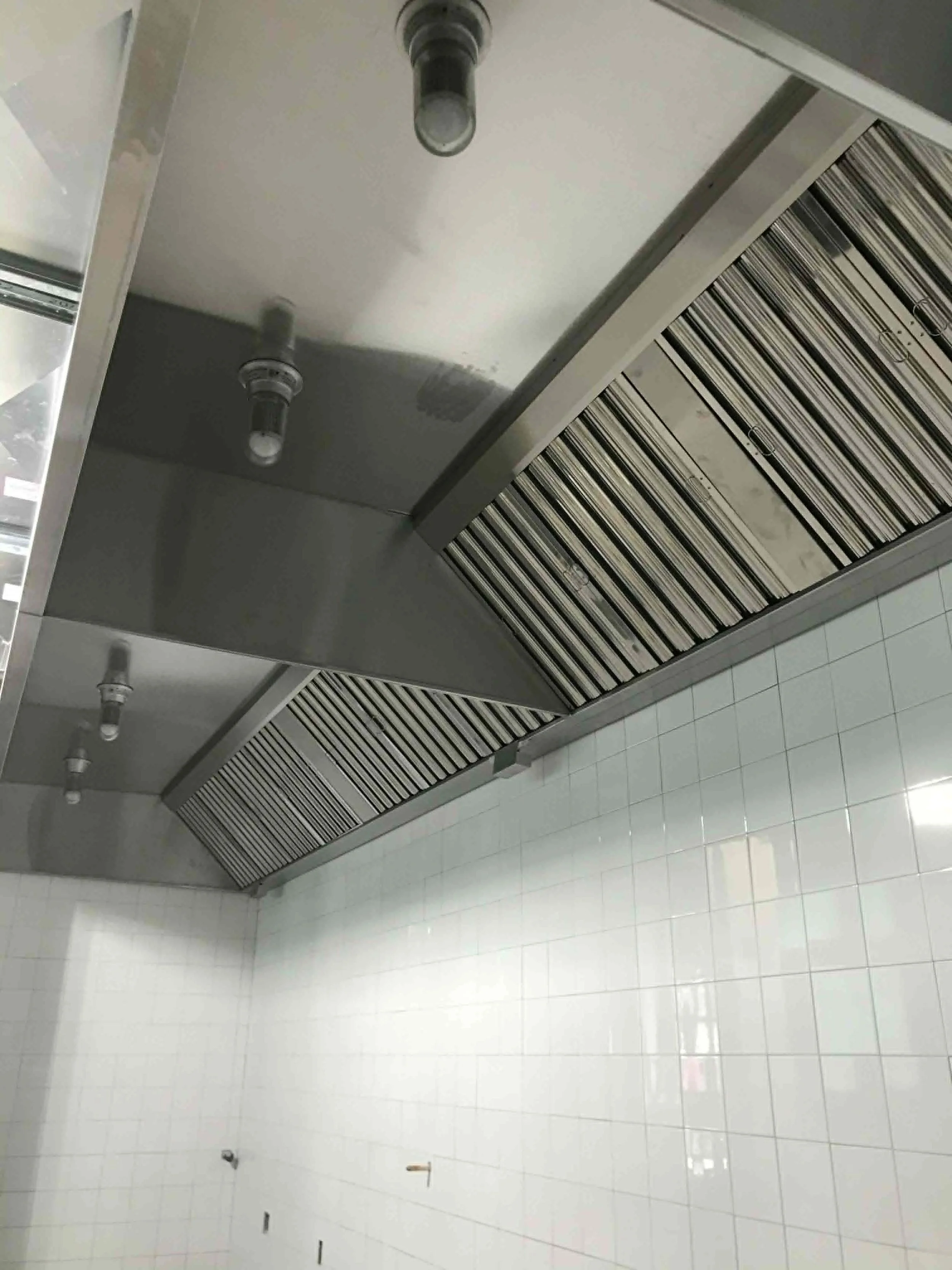 Une cuisine ou un espace commercial avec un plafond en métal qui cache des systèmes de ventilation. Les murs sont recouverts de carreaux blancs, et il y a des appareils d'éclairage suspendus. La pièce semble être en rénovation ou en construction.