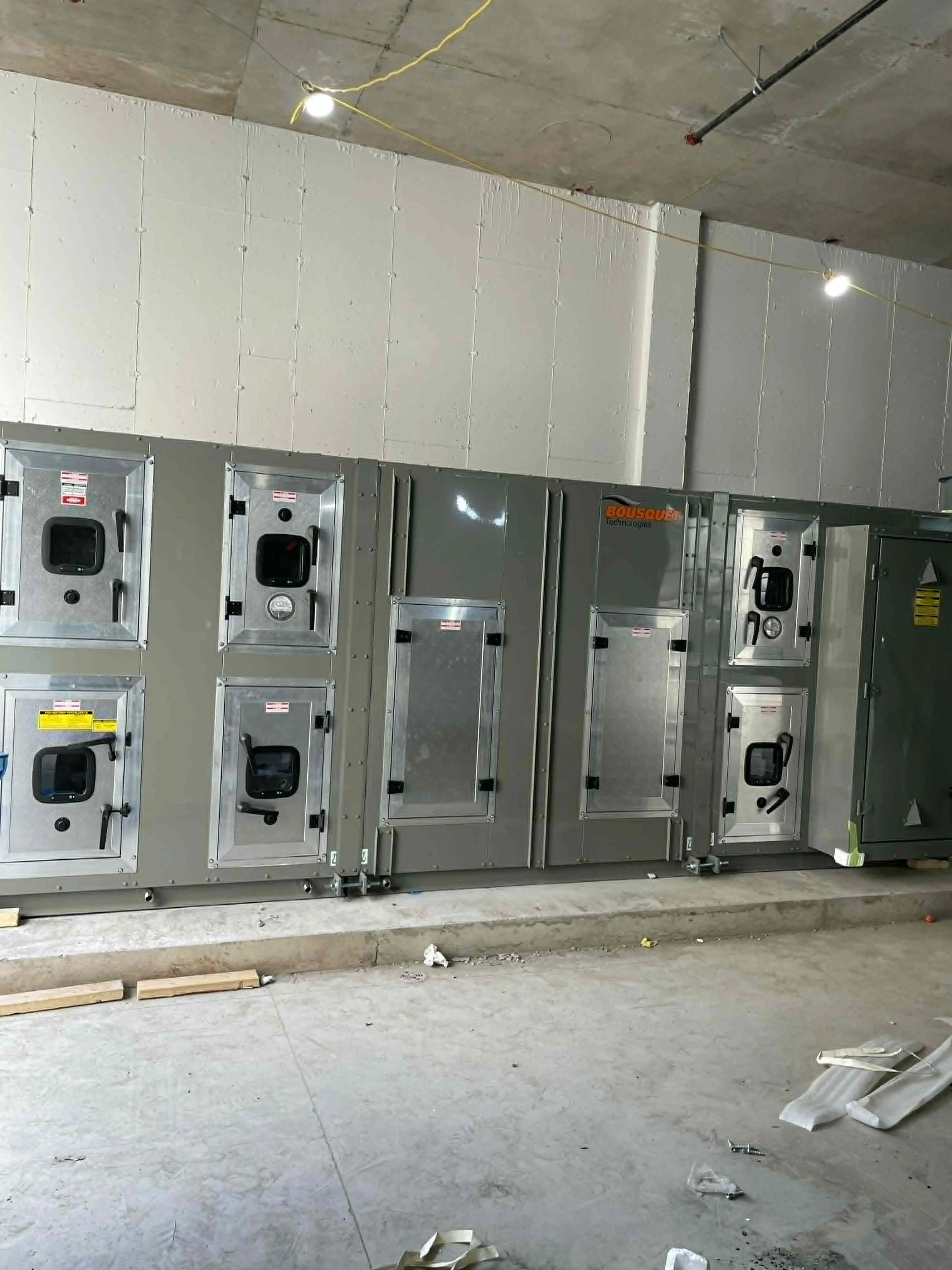 Installation électrique en cours avec des coffrets électriques métalliques alignés le long du mur, et des câbles électriques suspendus au plafond. Sol en béton avec des débris et matériaux de construction autour.