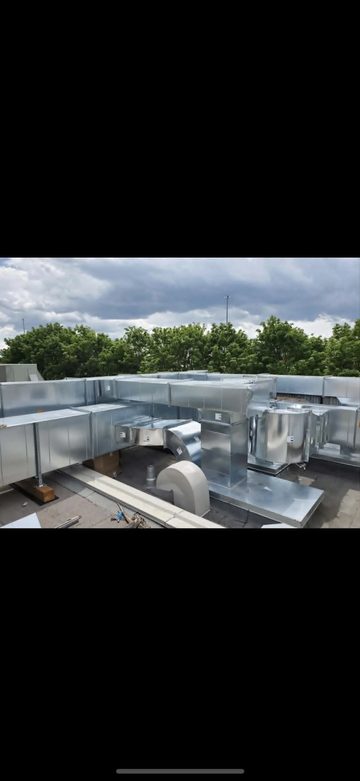 Vue d'une installation de conduits de ventilation métalliques sur un toit, avec des arbres et un ciel nuageux en arrière-plan.