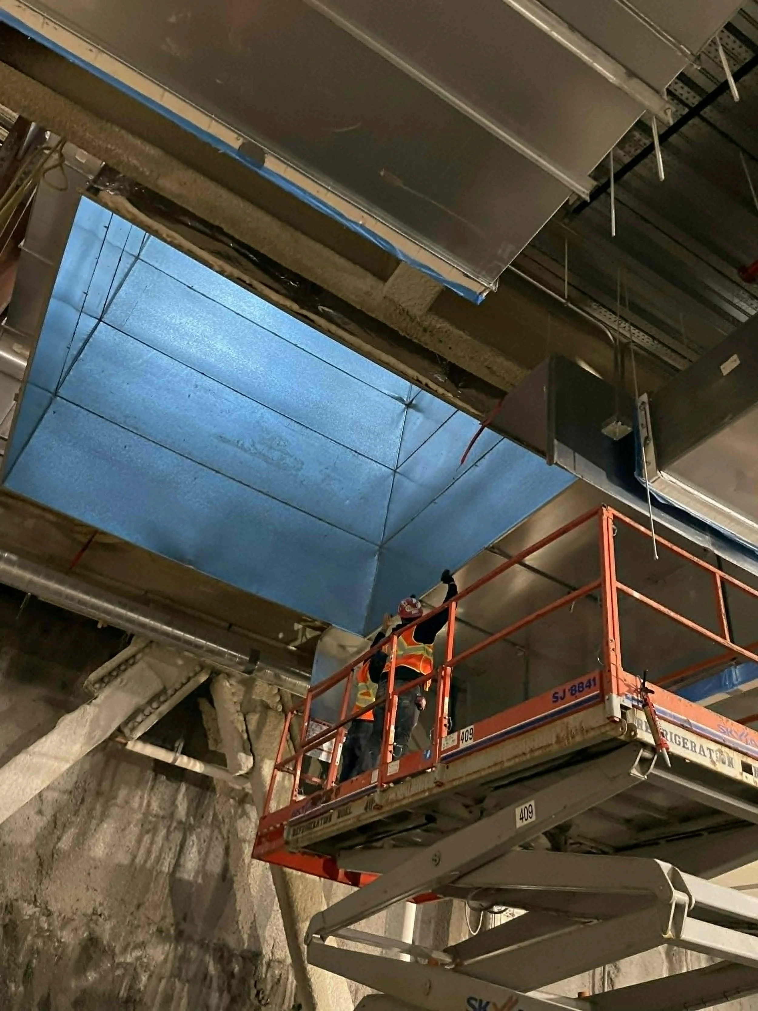 Techniciens travaillant sur une plateforme élévatrice en intérieur, installation ou réparation de conduits ou plafonds suspendus.