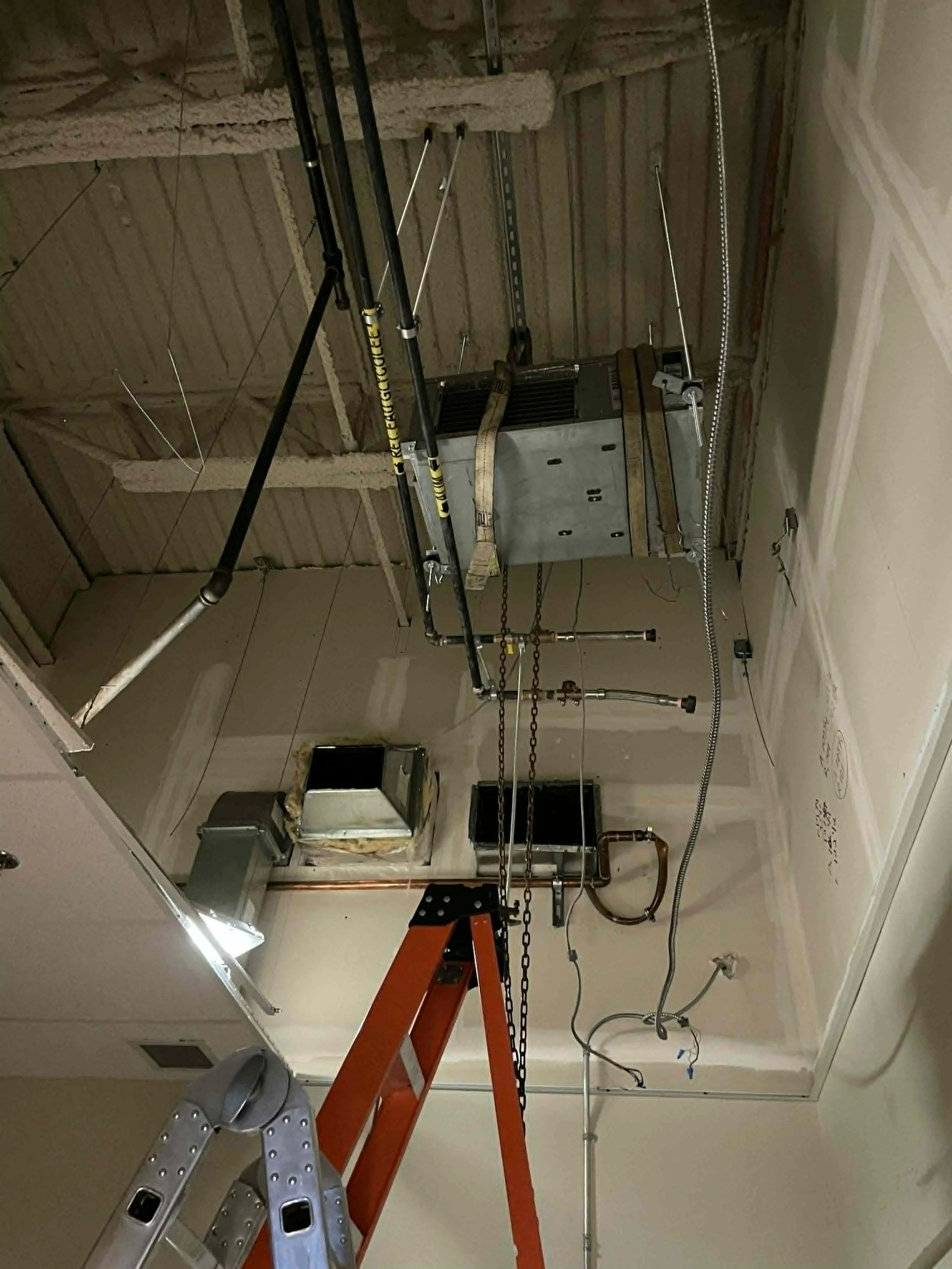 Construction en cours dans une pièce avec des murs en plâtre, un plafond en travaux, un escabeau orange, des tuyaux, des câbles, un circuit de ventilation et une lumière LED